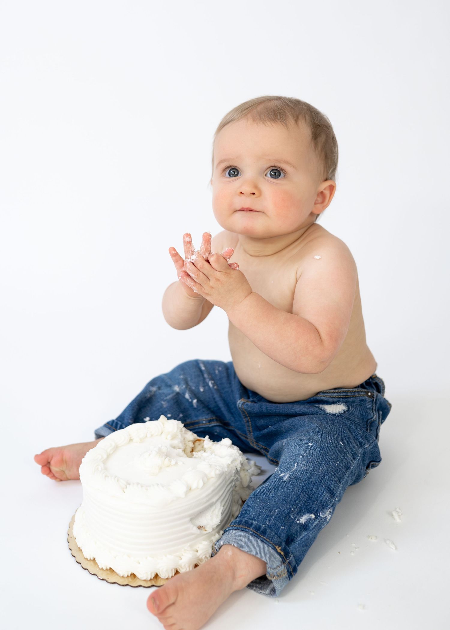 Baby in jeans clapping hands next to smashed white birthday cake on white background.
