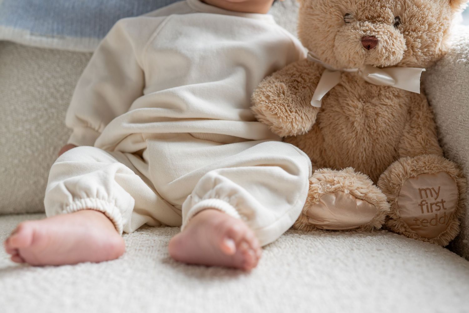 A baby's feet in white socks and booties rest next to a plush teddy bear on a soft surface.