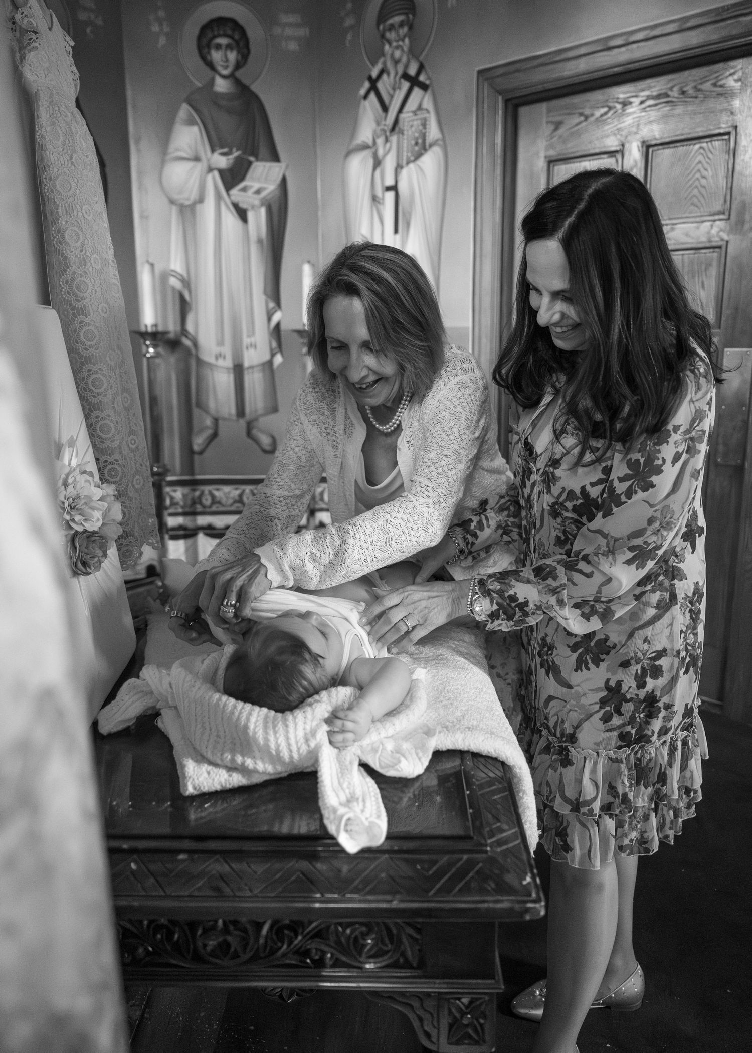 Two women in floral dresses attending to a baby on an ornate changing table in a room with religious icons.