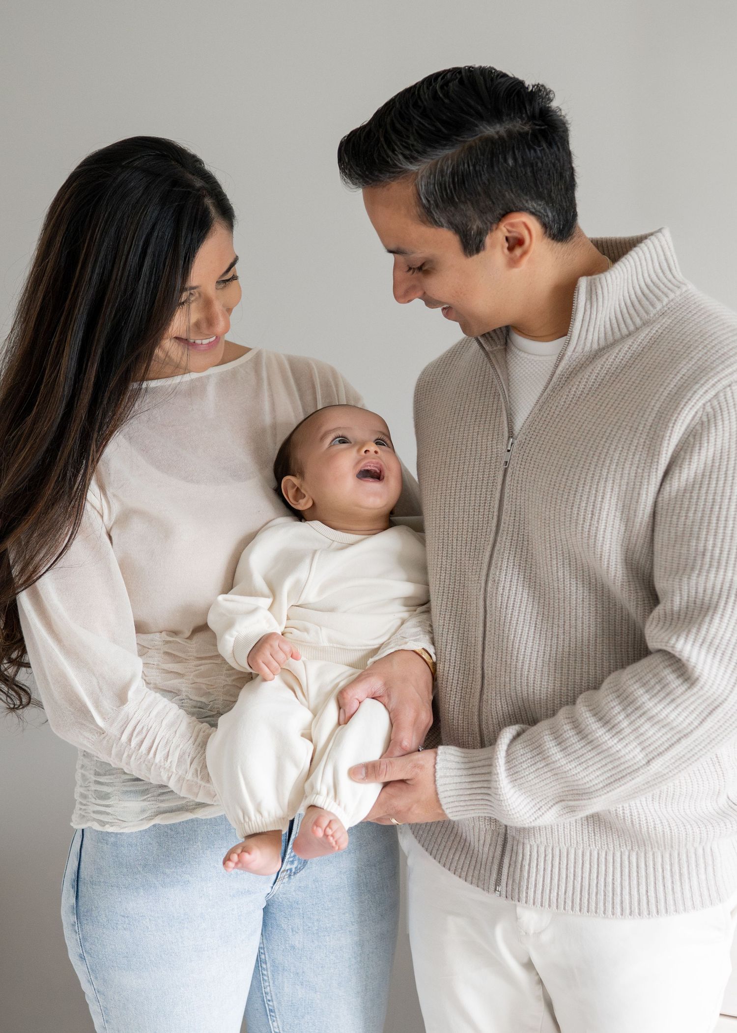 Happy young family with mother and father holding their smiling newborn baby in white outfit against gray background.