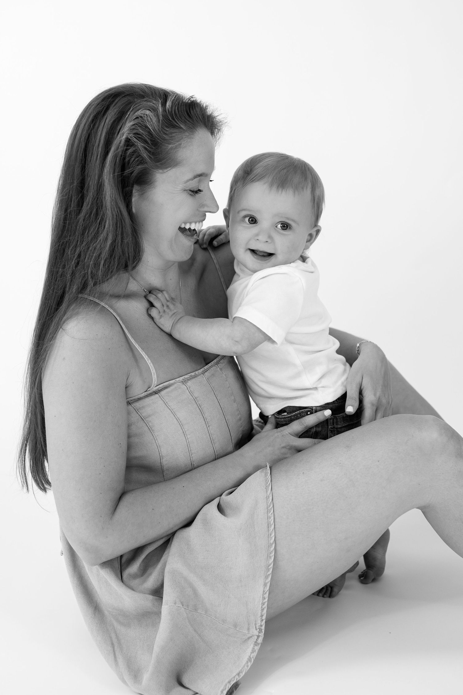 Black and white photo of a smiling mother sitting cross-legged holding her happy baby boy in a studio setting.