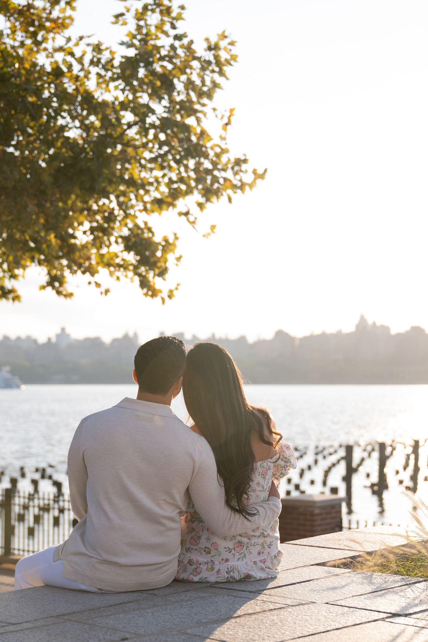 Couple sits together watching sunset by waterfront with wooden posts visible in background.