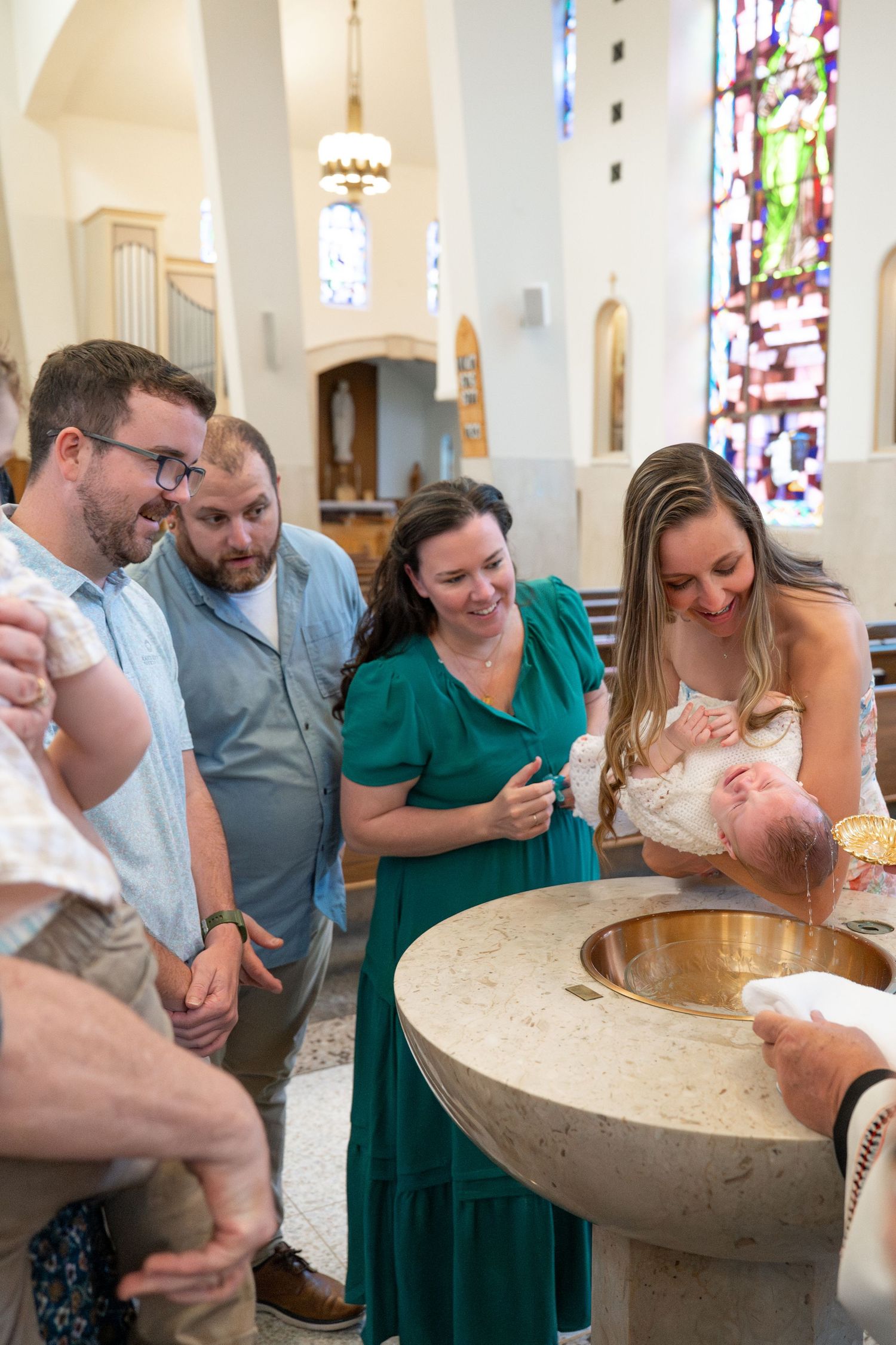 A group gathers around a baptismal font in a church with stained glass windows during a christening ceremony.