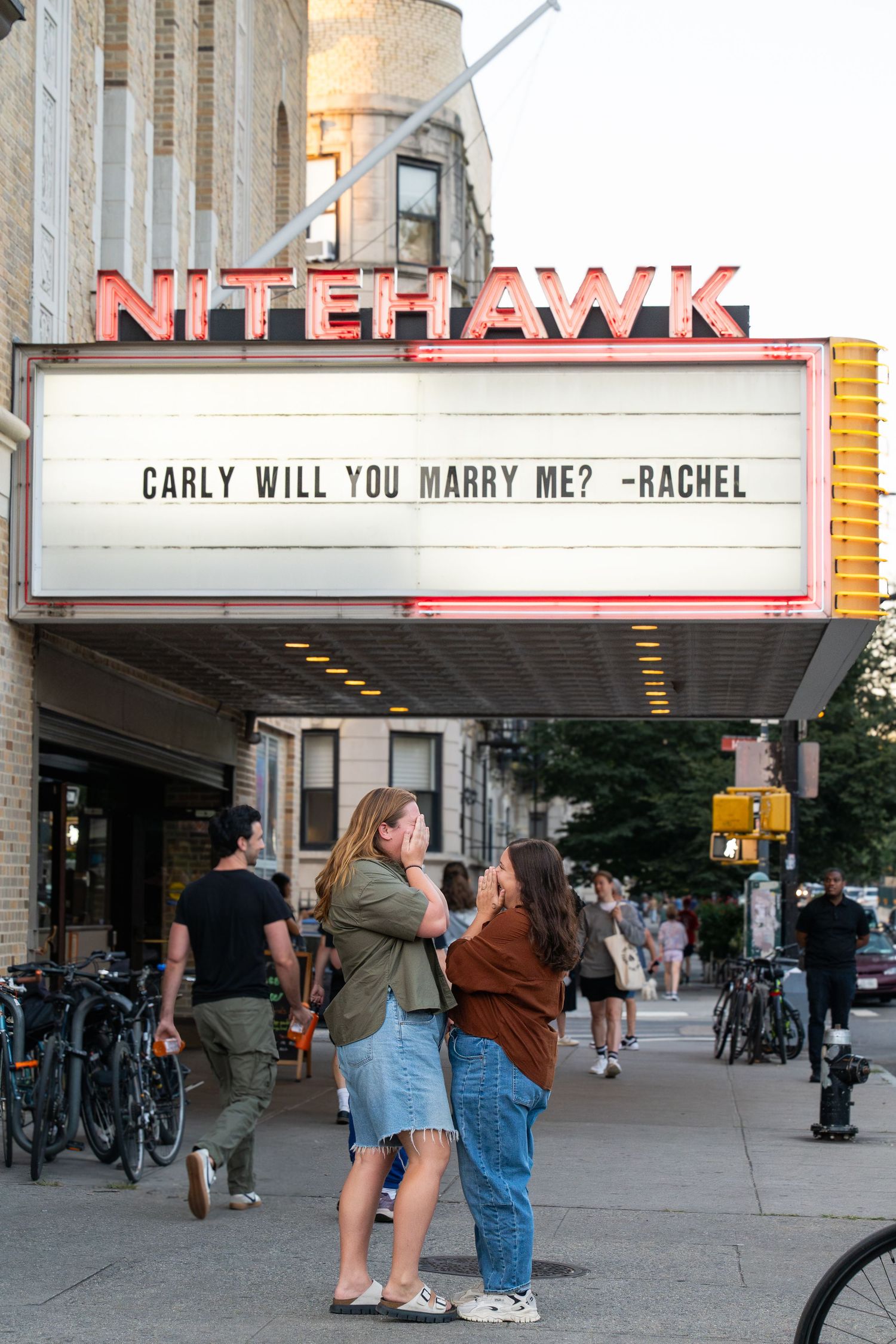 Movie theater marquee displays a marriage proposal reading 'CARLY WILL YOU MARRY ME? -RACHEL' in front of the Nitehawk Cinema.