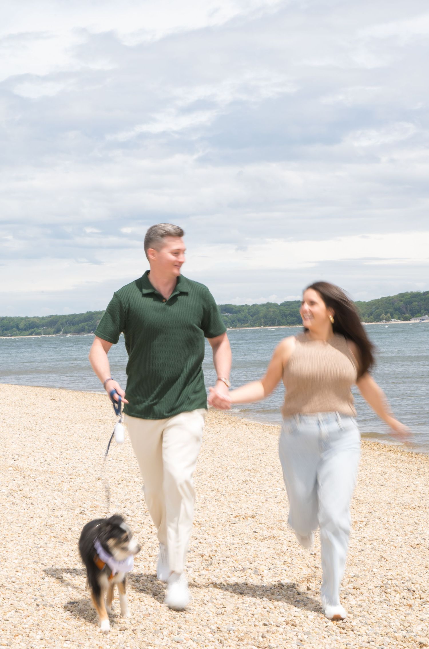 A couple walks their dog along a scenic beach on a cloudy day.