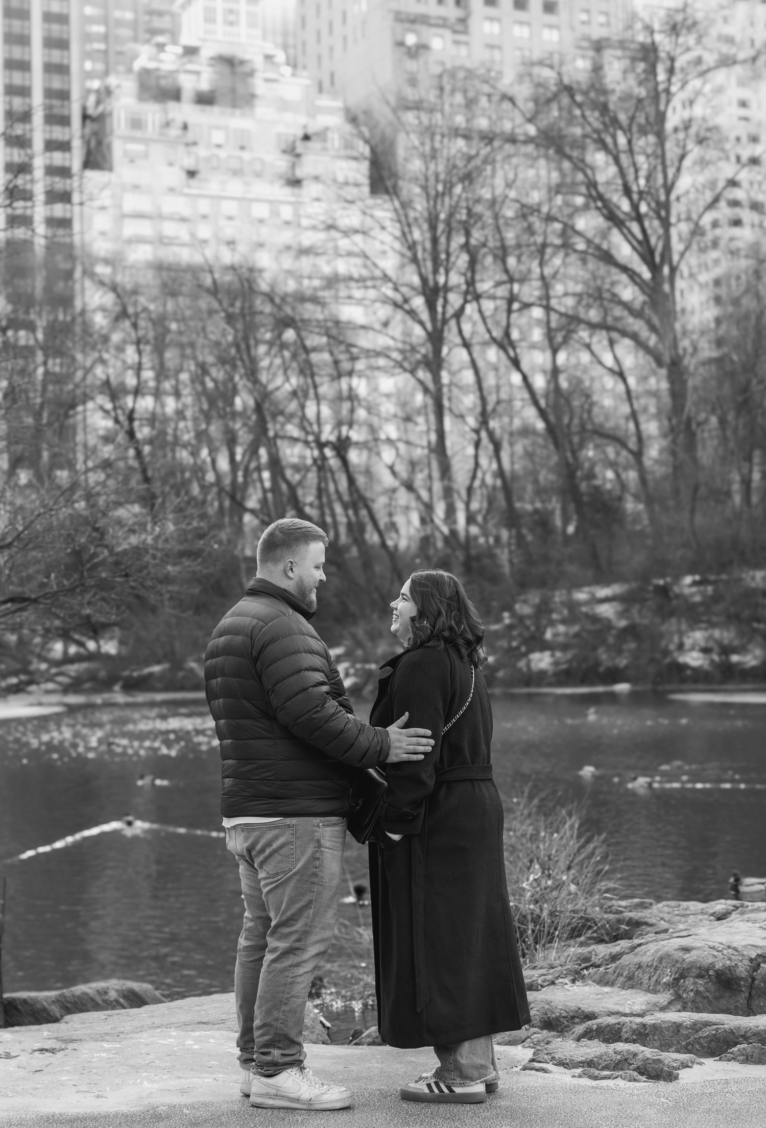 A romantic couple shares an intimate moment by a lake in Central Park with city buildings visible in the background.