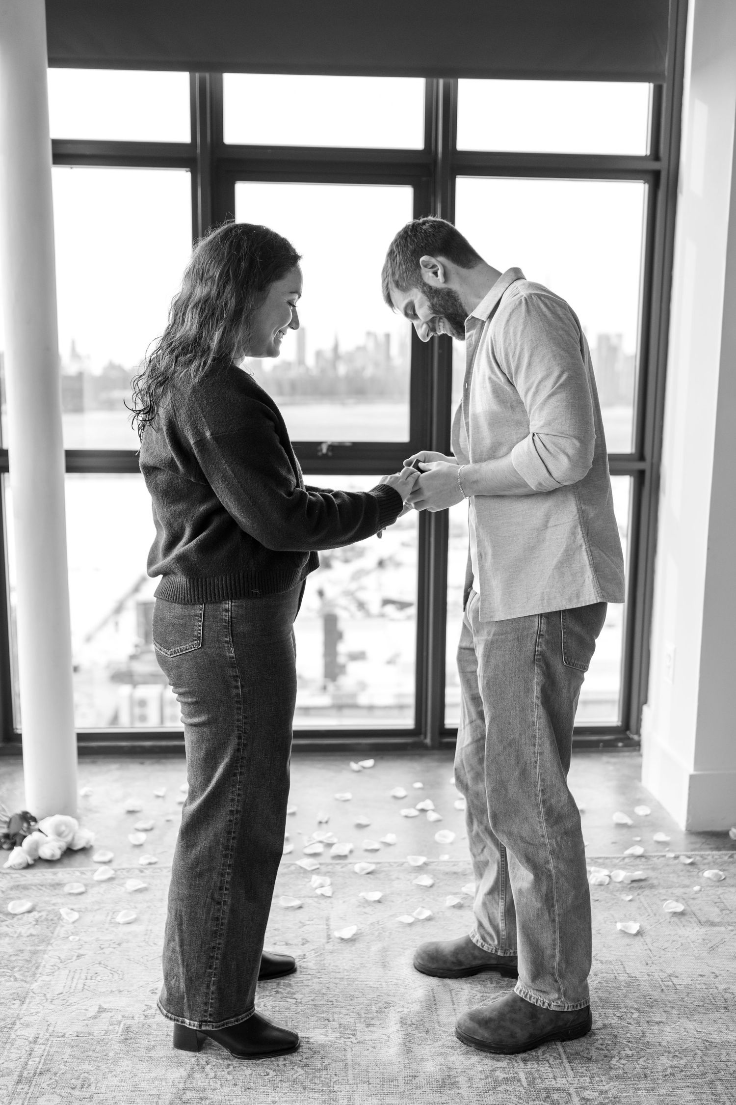 A couple shares a tender moment in black and white by a window overlooking a city skyline.