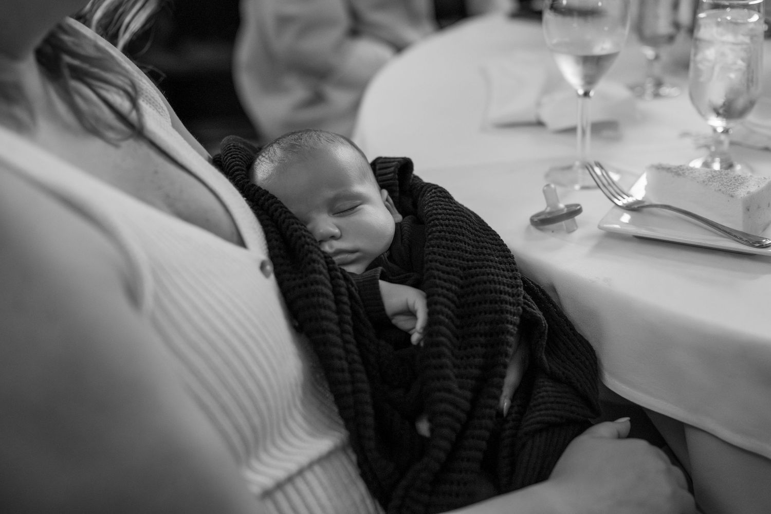 A sleeping baby wrapped in a black sweater rests at a formal dining table setting.
