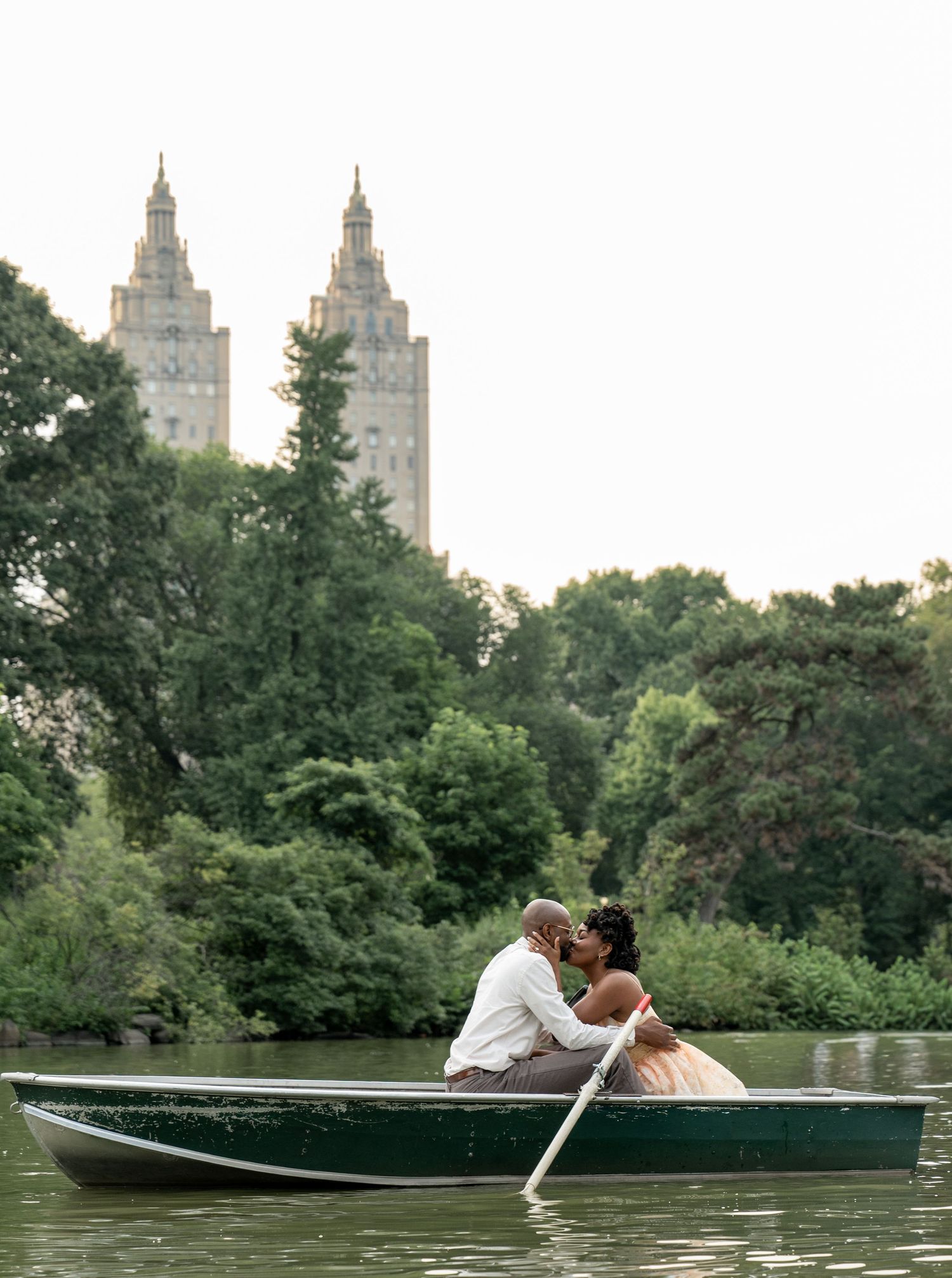 Romantic couple sharing tender moments in a rowboat on Central Park lake with iconic buildings in background.