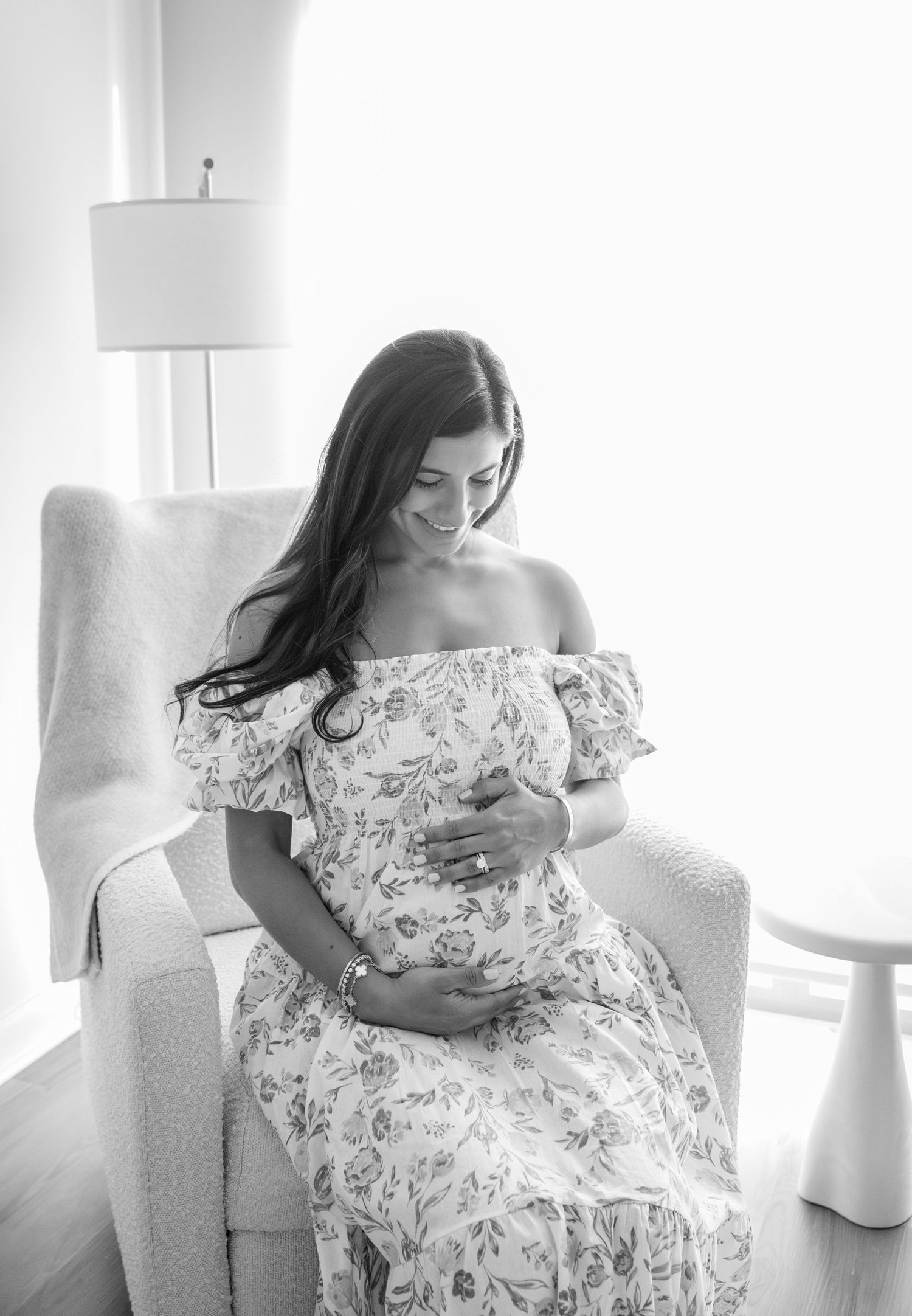 Woman in floral dress poses on white chair in bright interior setting.