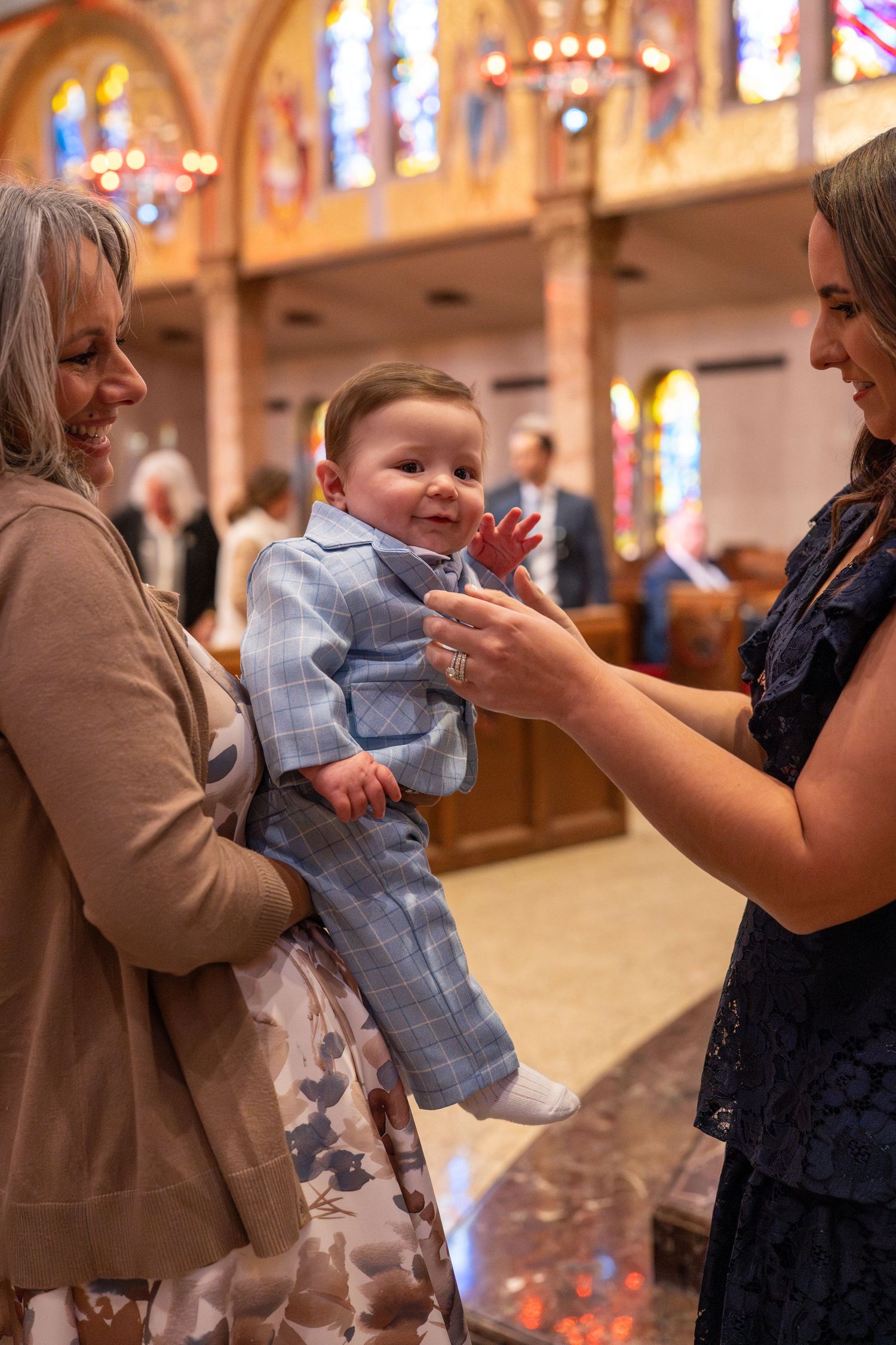 A baby in a light blue outfit is held during a church ceremony with stained glass windows in the background.