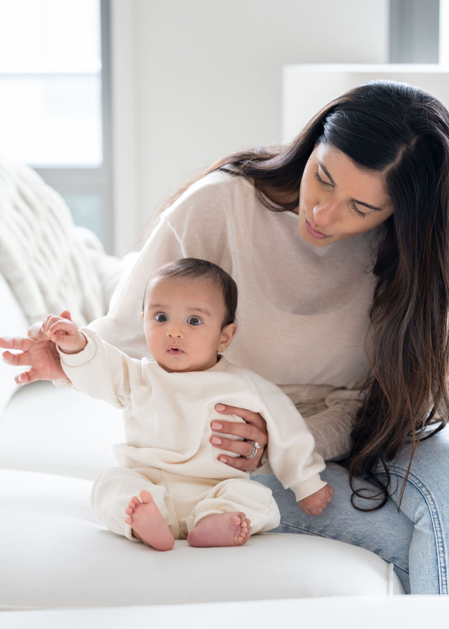 Dark-haired mother in beige sweater supporting curious baby in cream romper on white sofa.