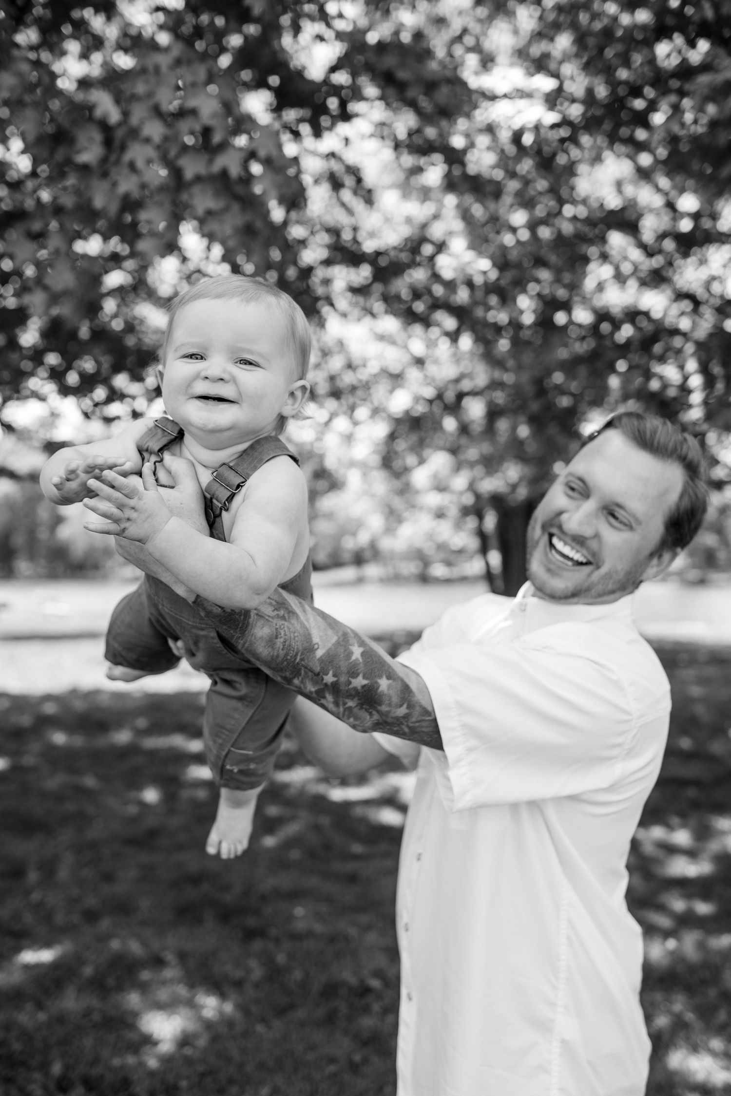 Black and white photo of laughing father lifting smiling baby in overalls outdoors in sunny park.