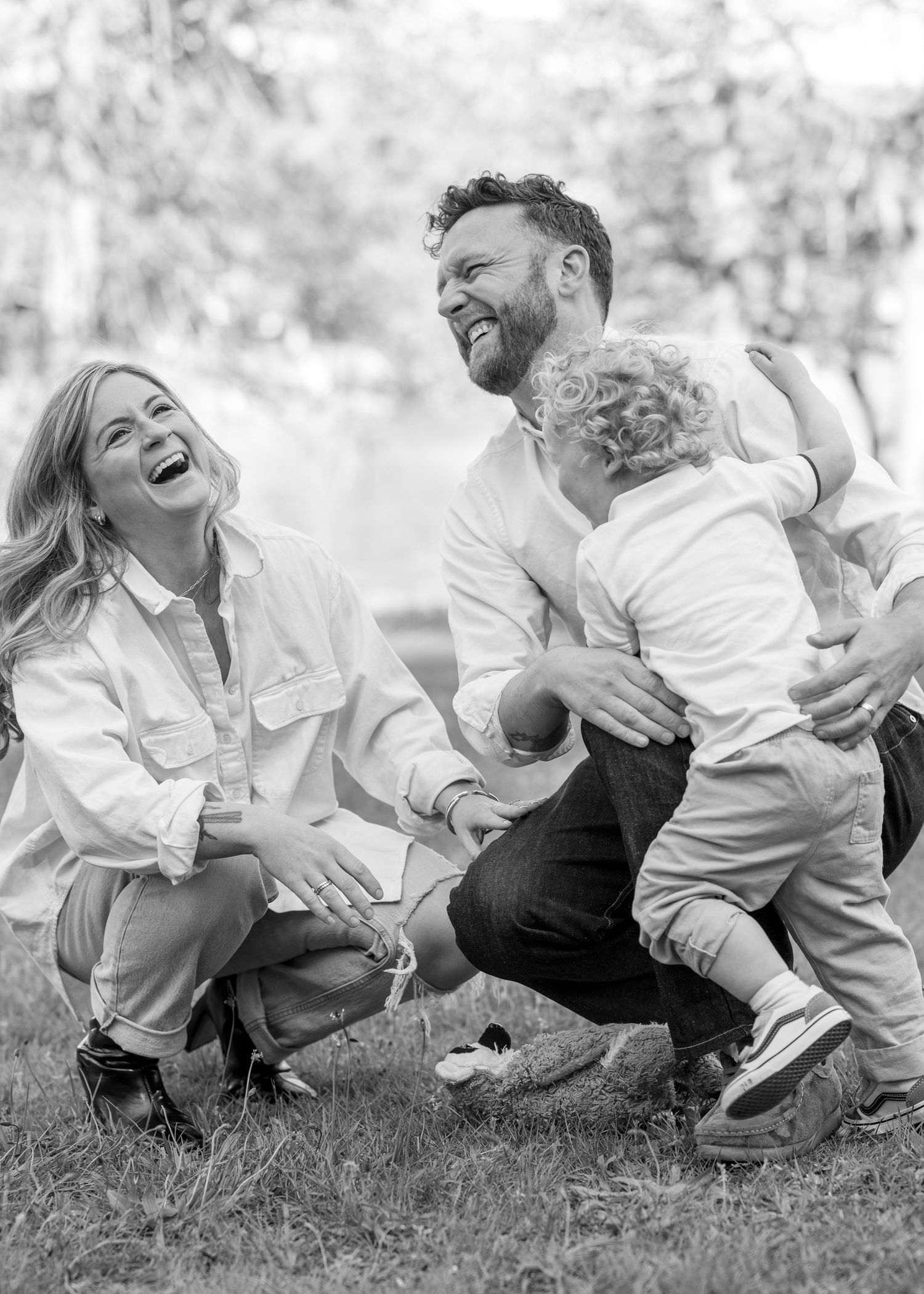 Black and white photo of a laughing family with a toddler playing outdoors in a park.