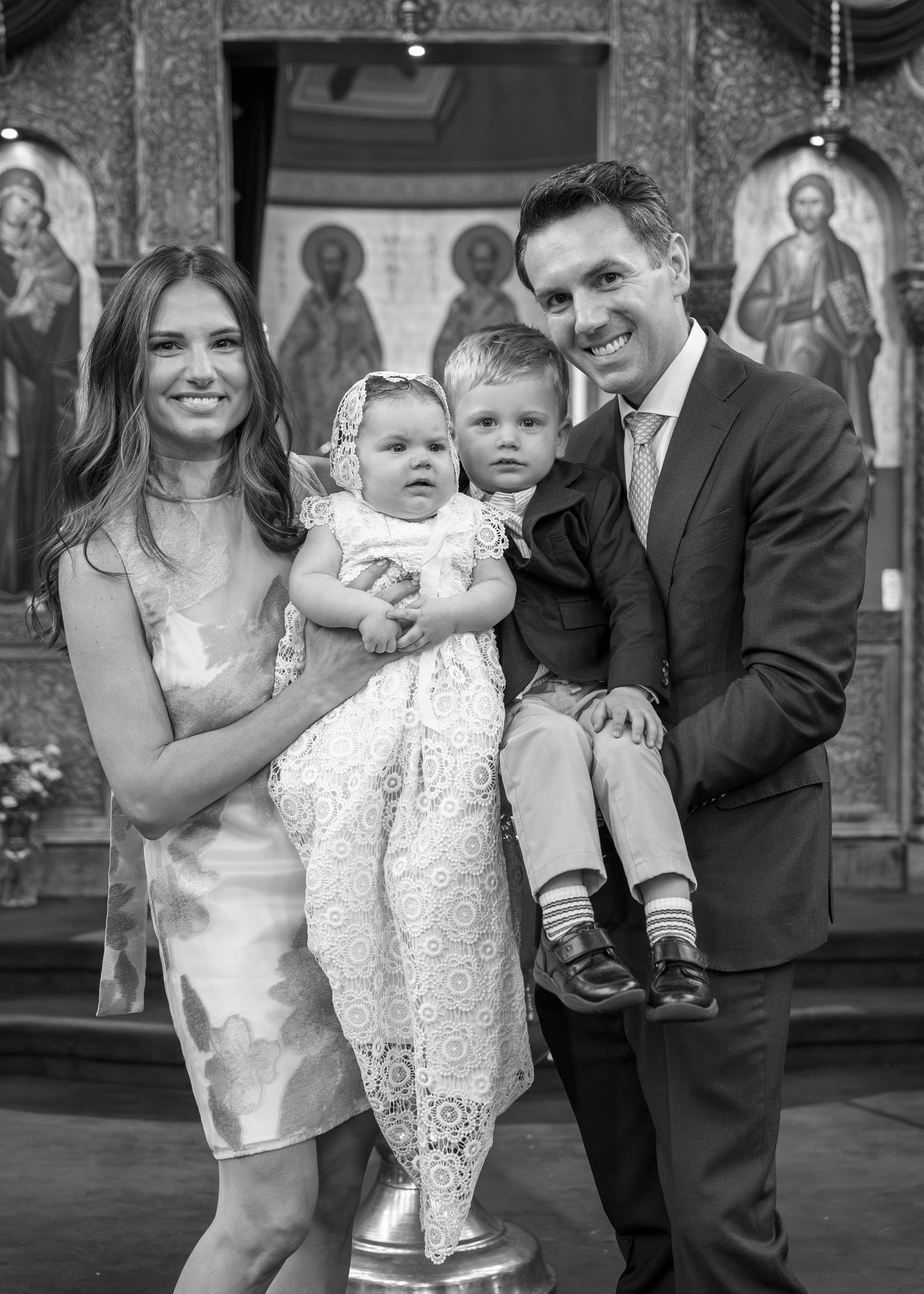 Black and white family portrait at a church baptism with parents holding two young children before religious icons.