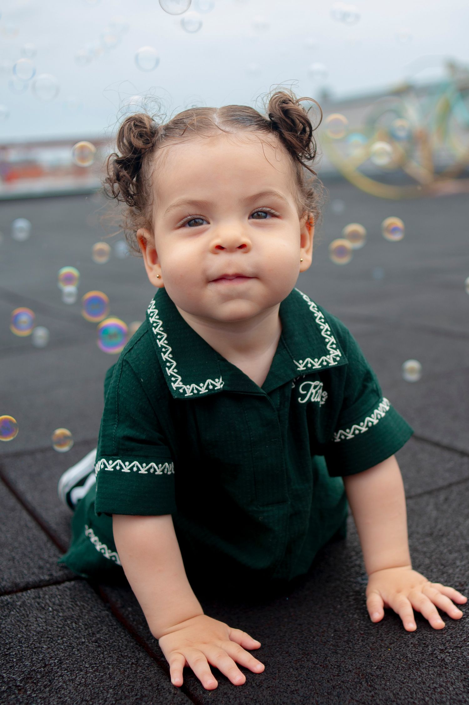 Baby in green outfit playing with soap bubbles on dark pavement.