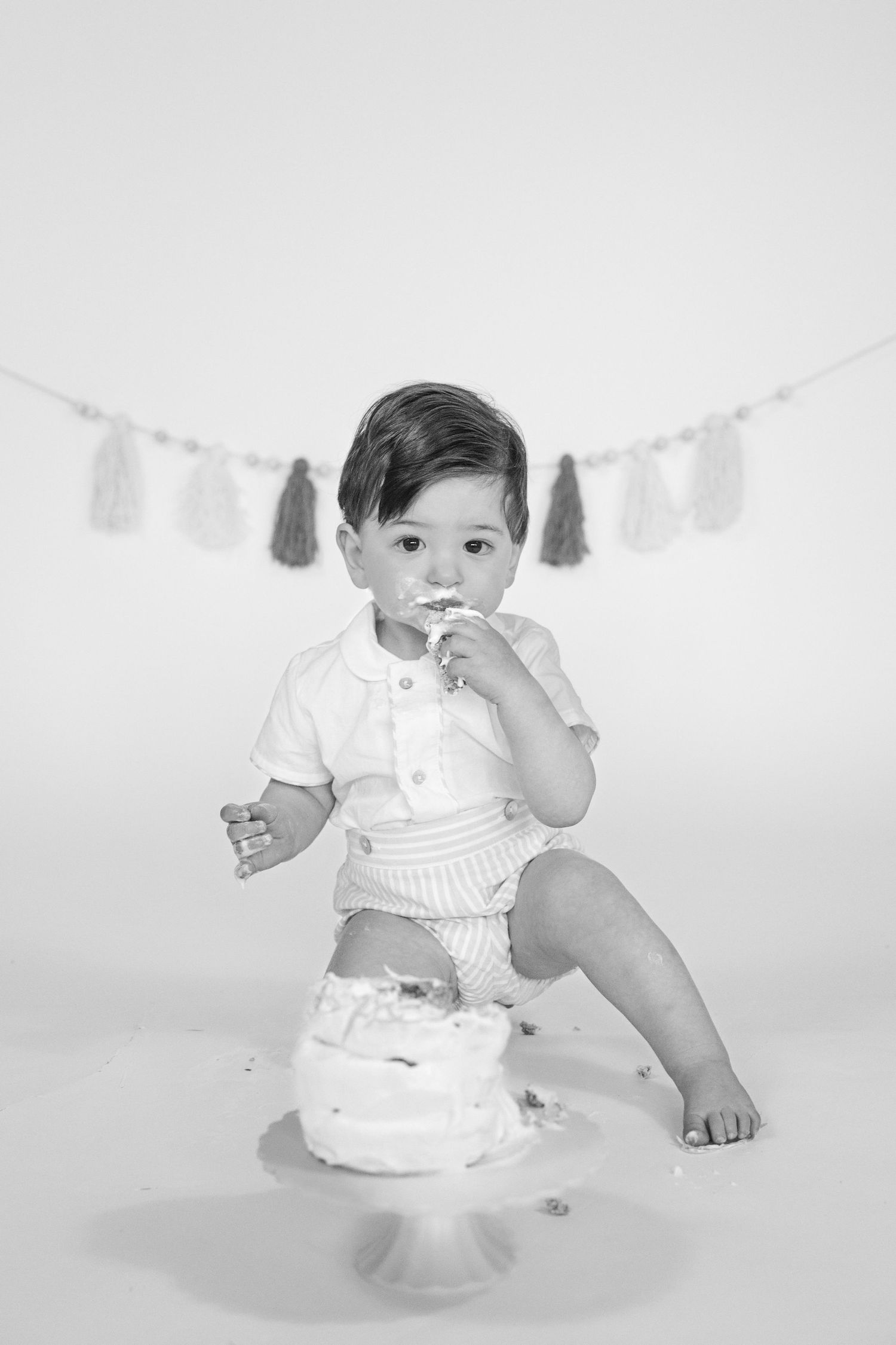 Baby in white outfit plays with cake while yellow and white tassels hang in background.