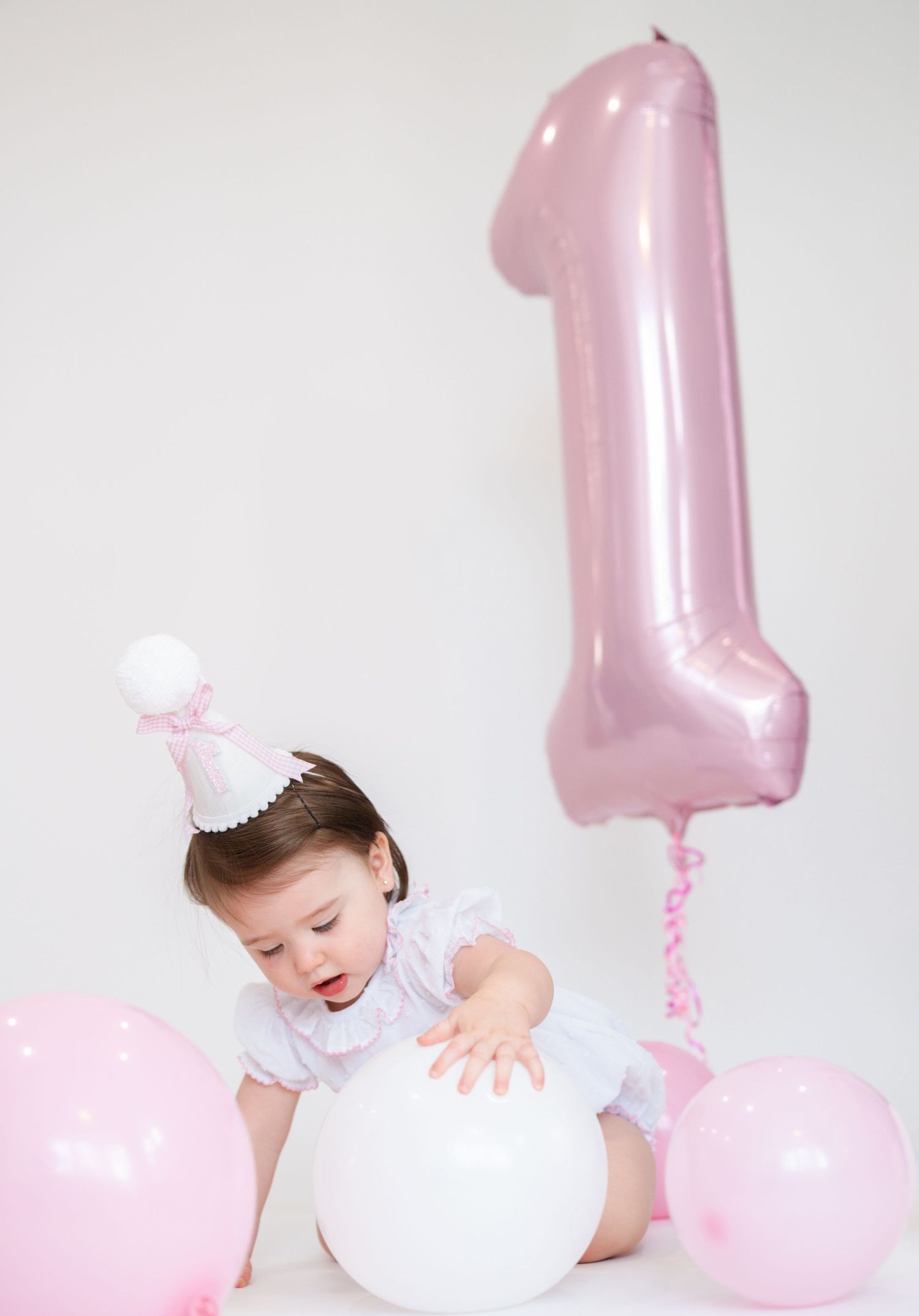 Baby in white outfit plays with pink and white balloons against white backdrop.
