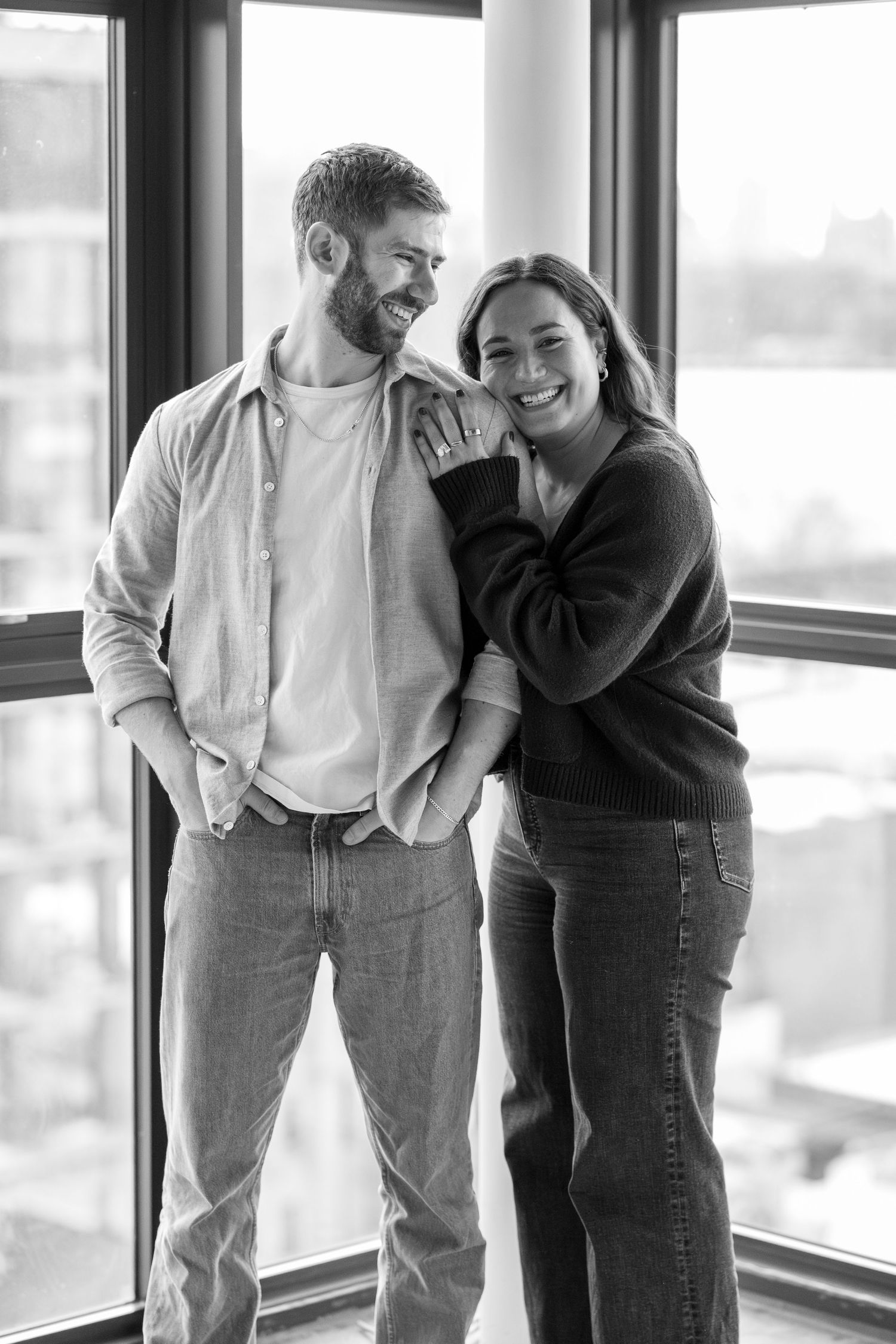 A couple shares candid laughter and embraces by large windows in a black and white photo series.