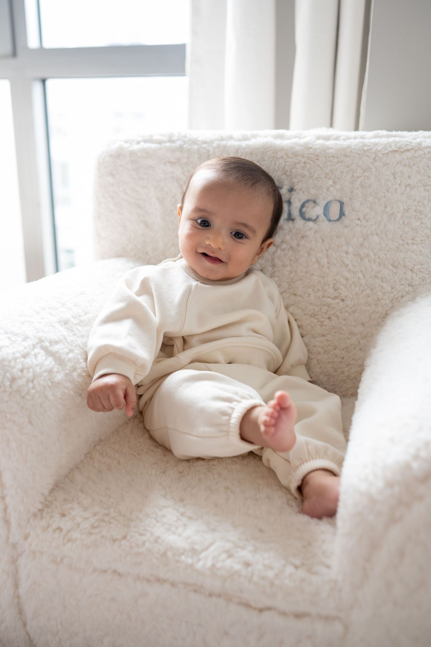 A smiling baby in cream-colored clothing lies on a plush white couch in a bright, sunlit room.