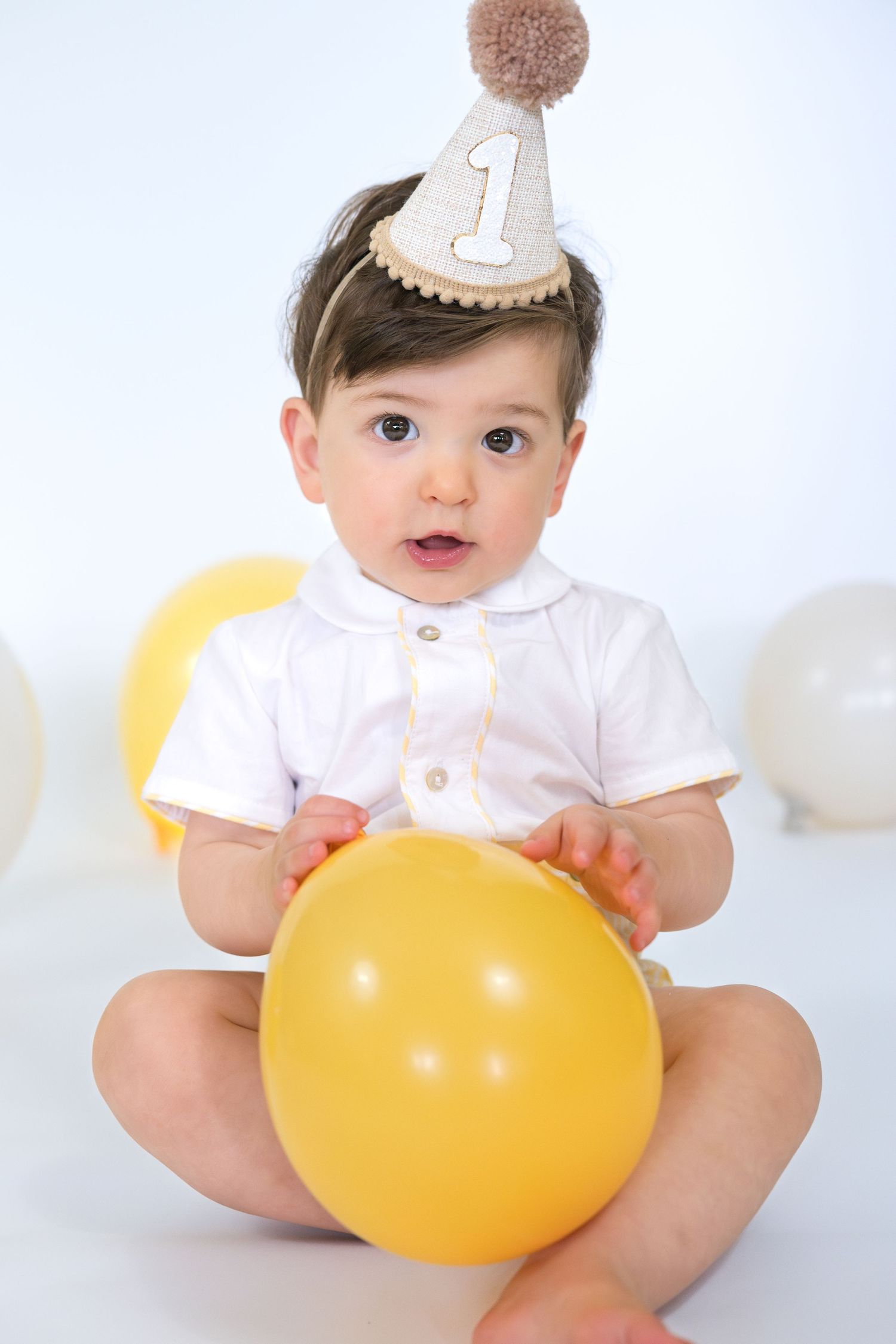Baby wearing party hat and white outfit holding yellow balloon during first birthday photoshoot.