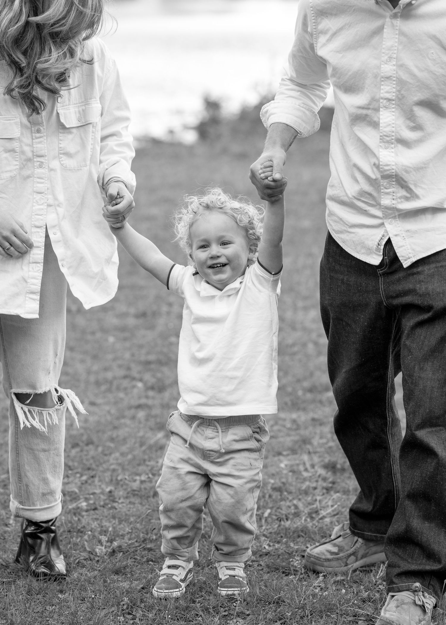 Black and white photo of a happy toddler holding hands with parents while walking outdoors on grass.