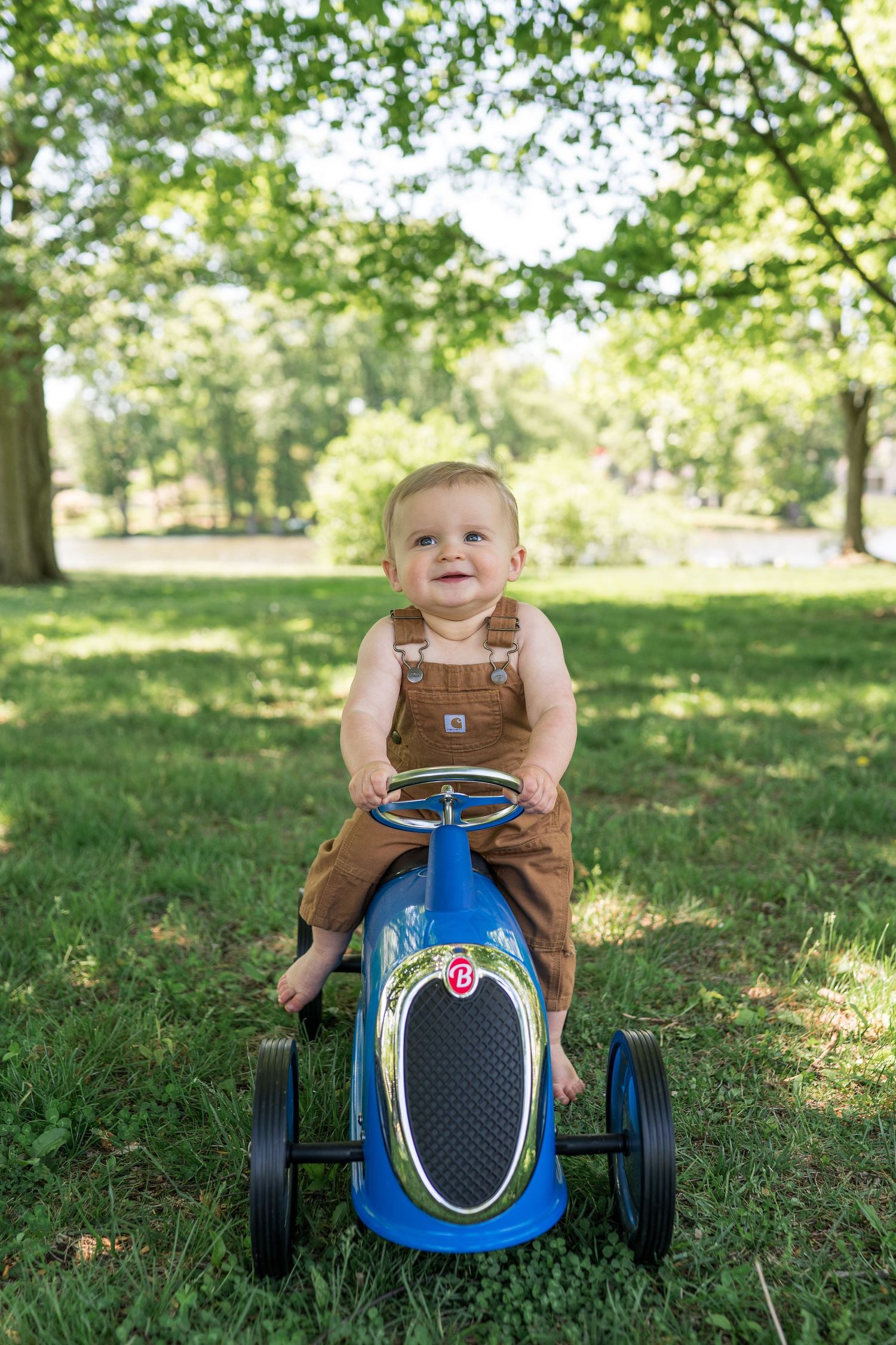 Smiling baby in brown overalls riding a blue vintage-style pedal car on green grass in a sunny park.
