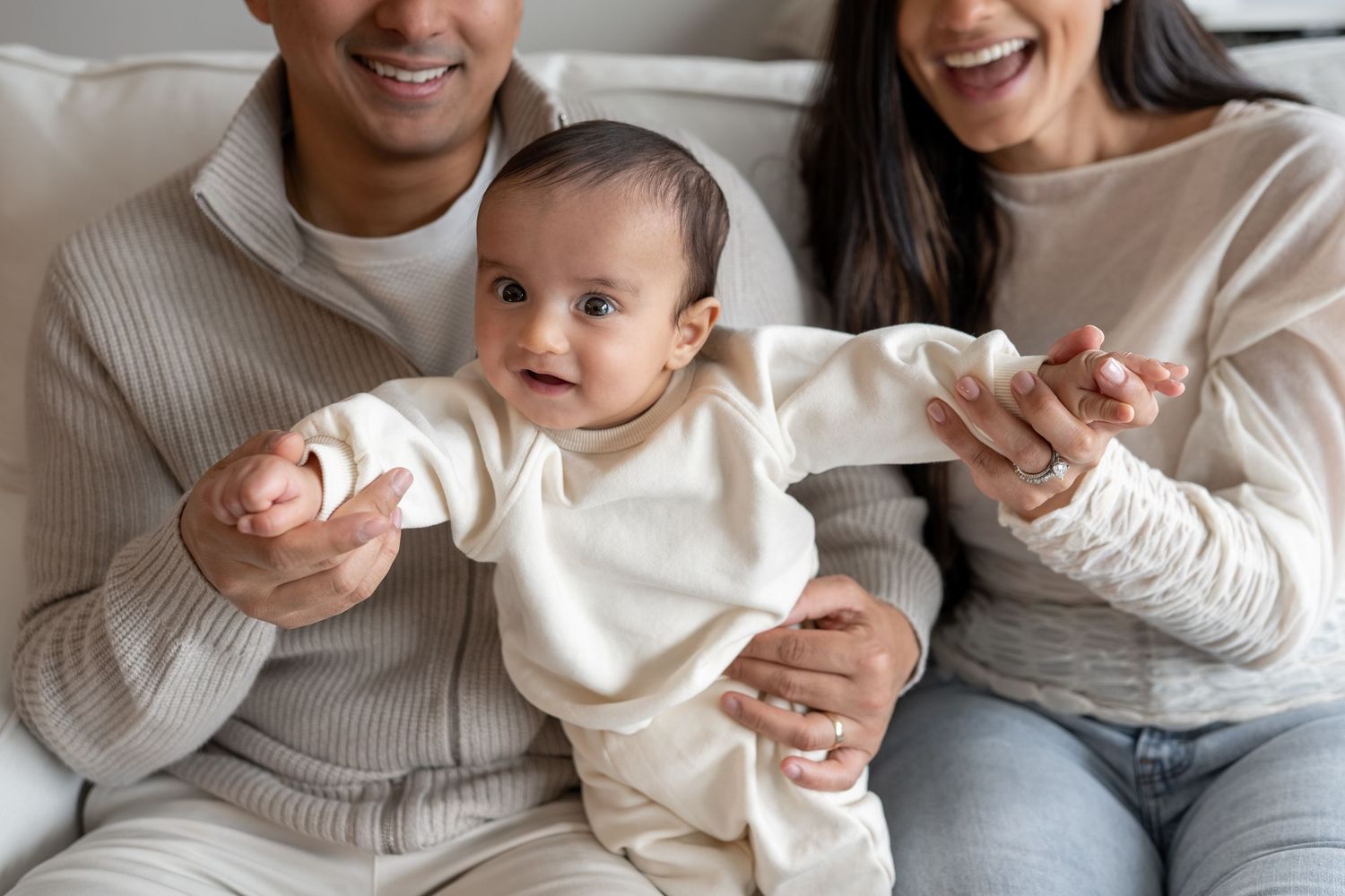 Smiling parents sitting on sofa with curious baby in cream outfit between them in bright living room.