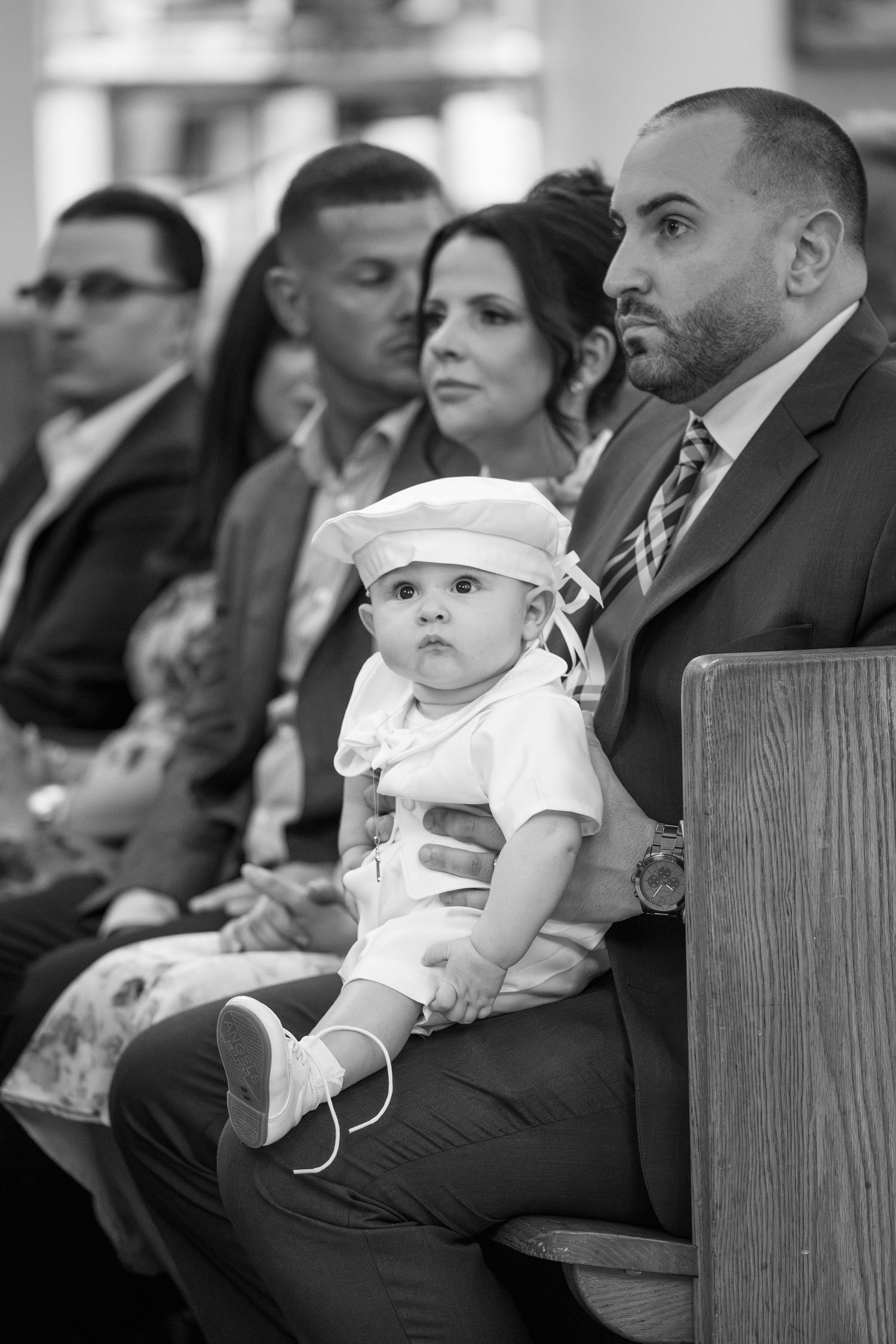 A baby in white formal wear sits on someone's lap during what appears to be a formal event.