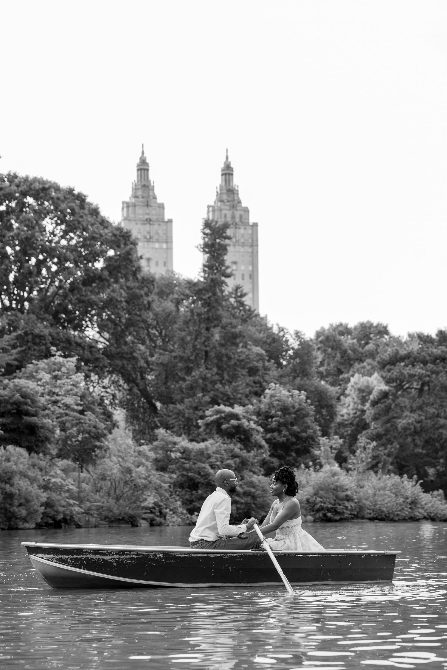 A rowboat glides across a tranquil lake with city buildings visible through the trees in the background.