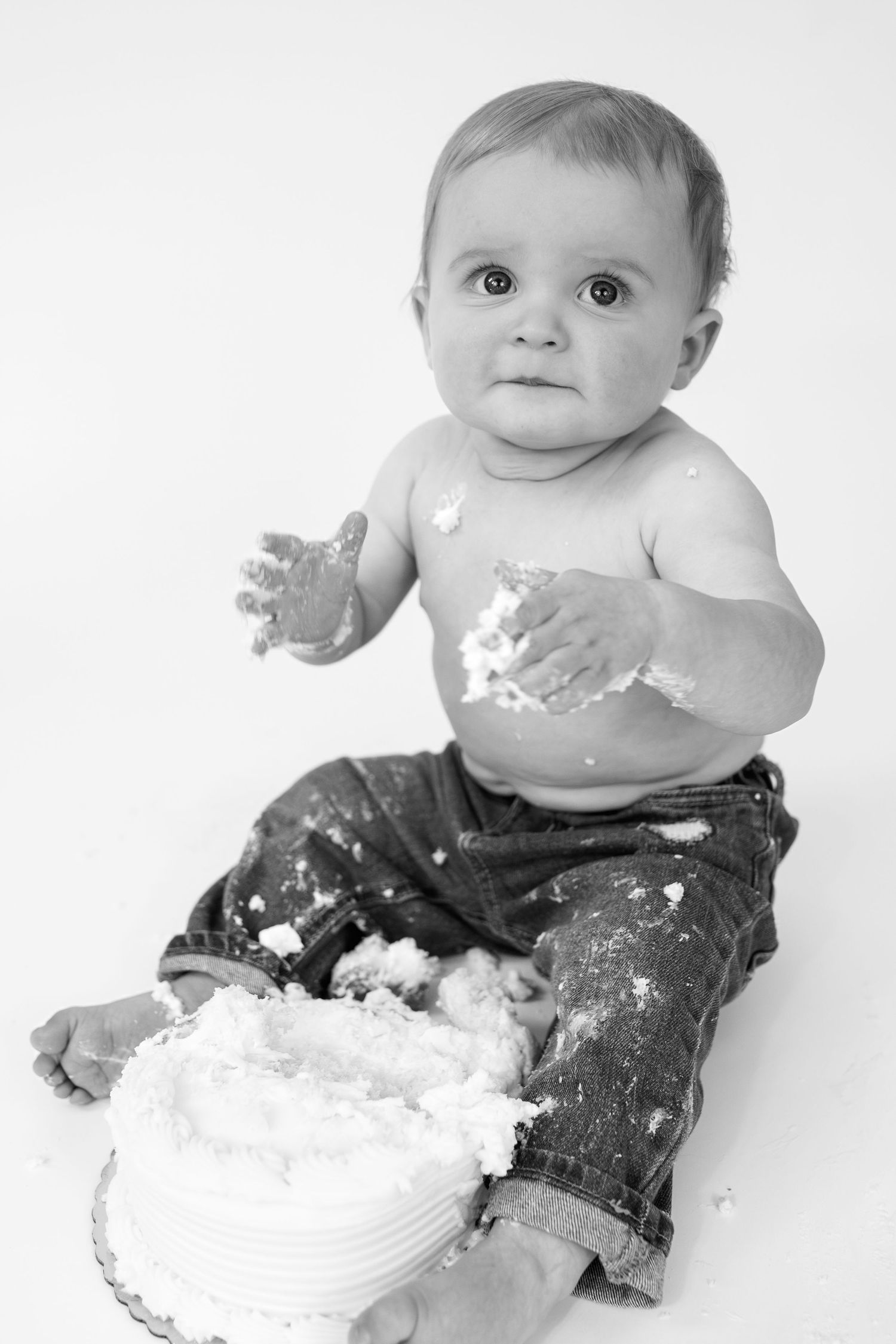 Black and white photo of a baby smashing a white cake, covered in frosting, wearing jeans, looking at camera.