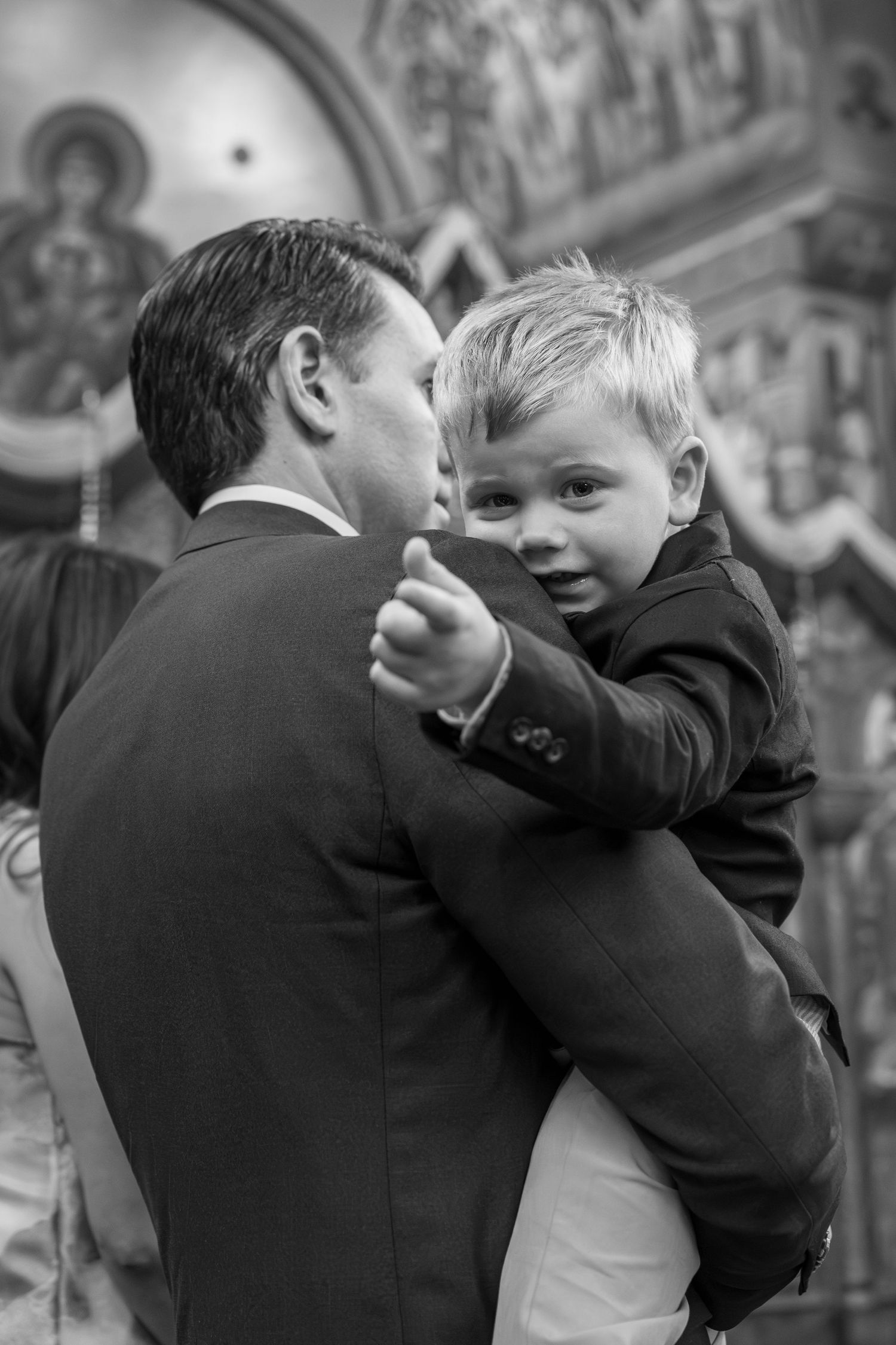 Young boy in suit smiling at camera while being held by man in formal attire inside ornate church during ceremony.