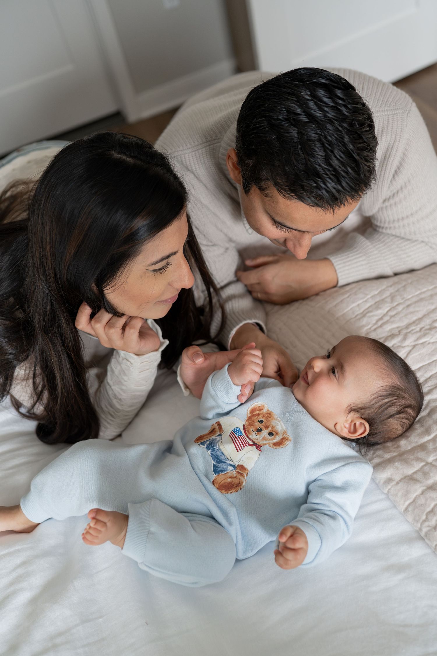 Parents lying on bed smiling at baby in blue bear onesie.