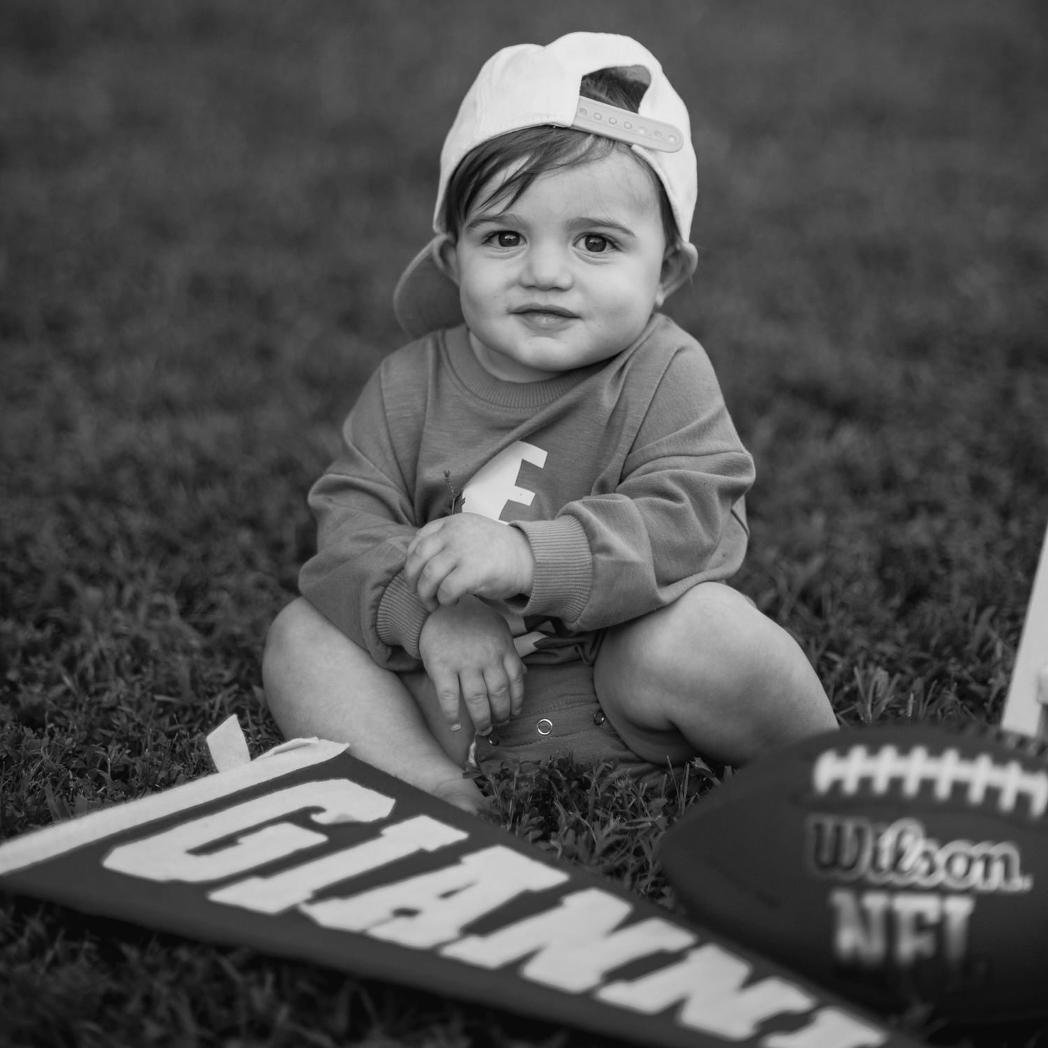 Baby in a baseball cap lying on grass with football equipment and text visible.