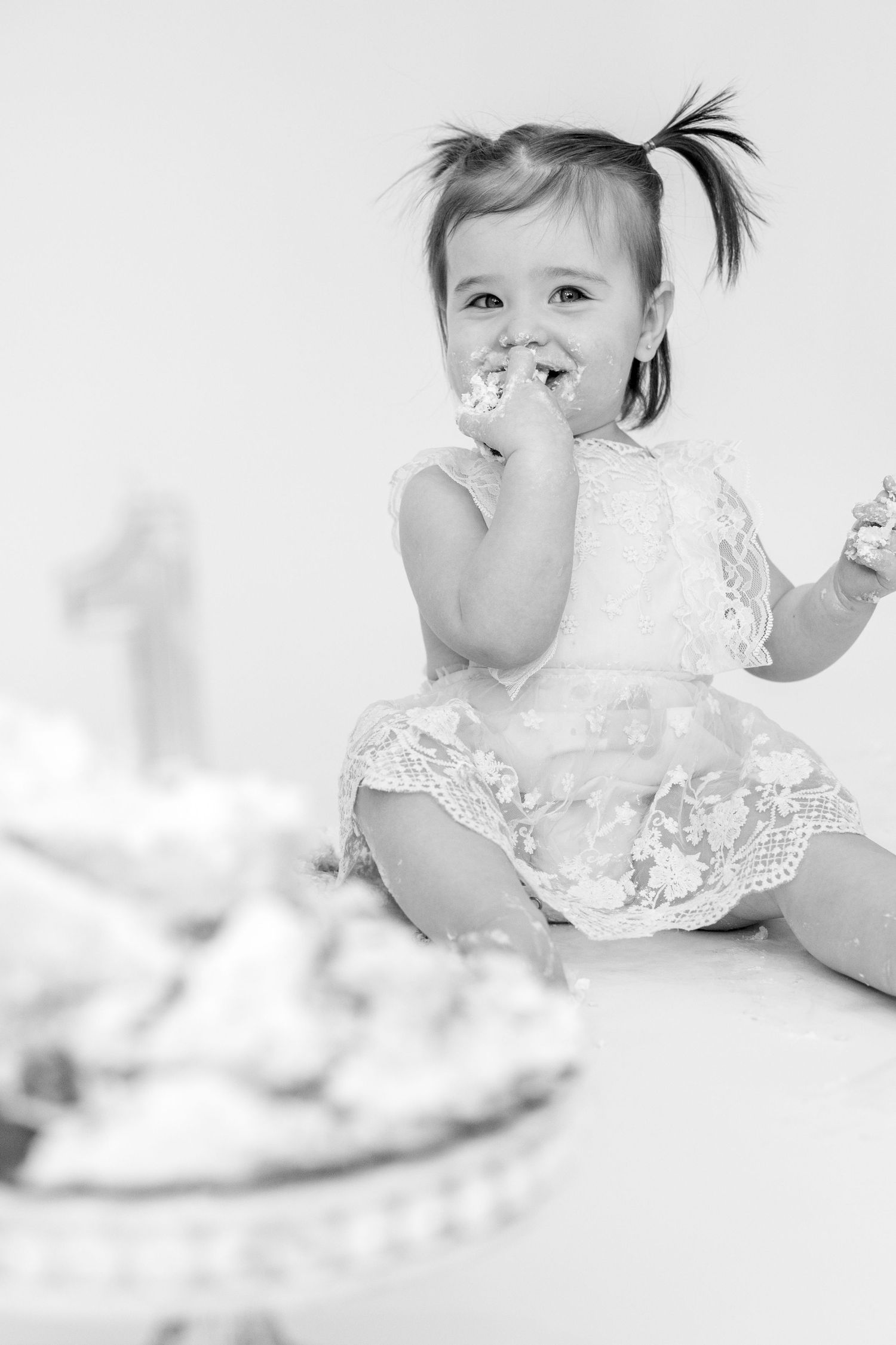 Black and white photo of toddler in lace dress enjoying cake.