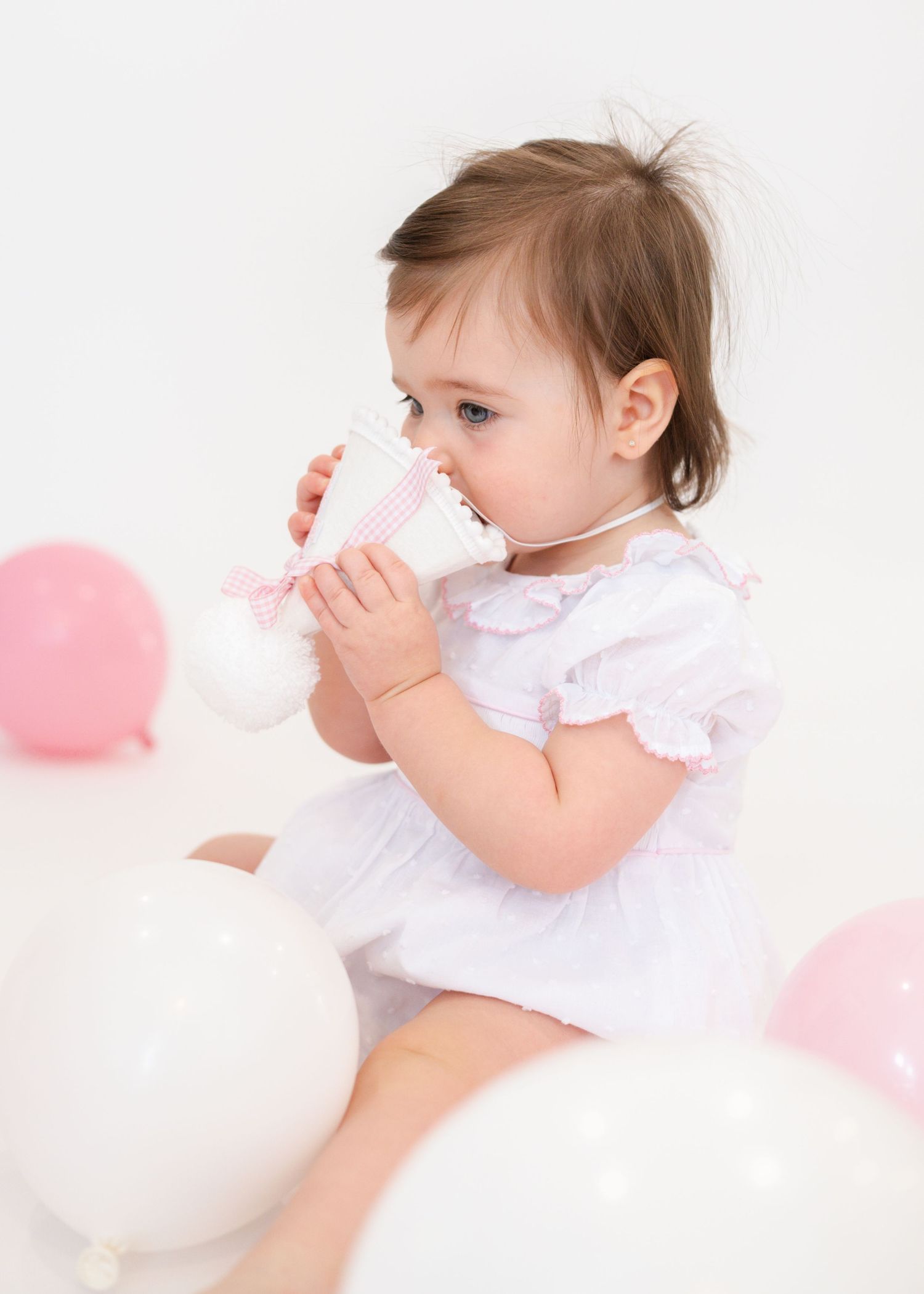 Baby in white ruffled dress playing with pink balloons during photo session.