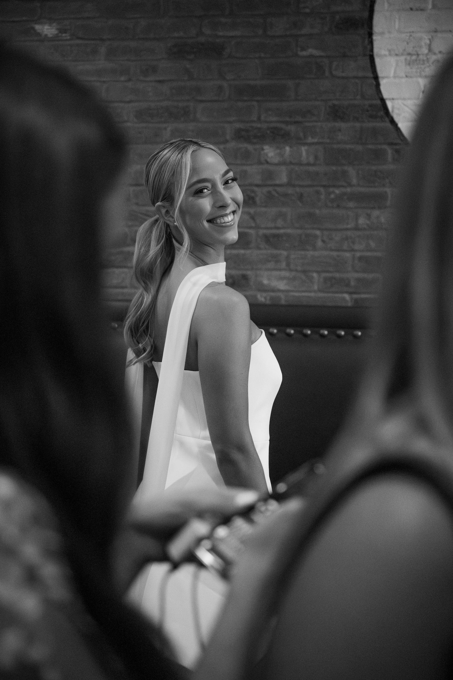 A candid black and white moment showing a person in a white dress smiling against a brick wall background.