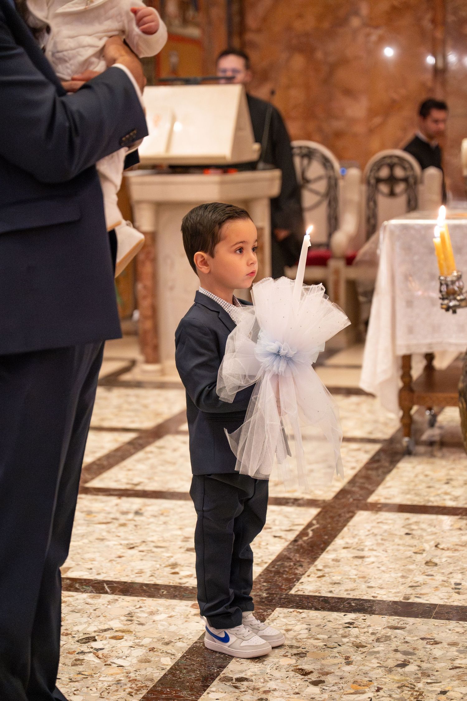 A young boy in a navy suit holds a decorated white candle during a church ceremony.
