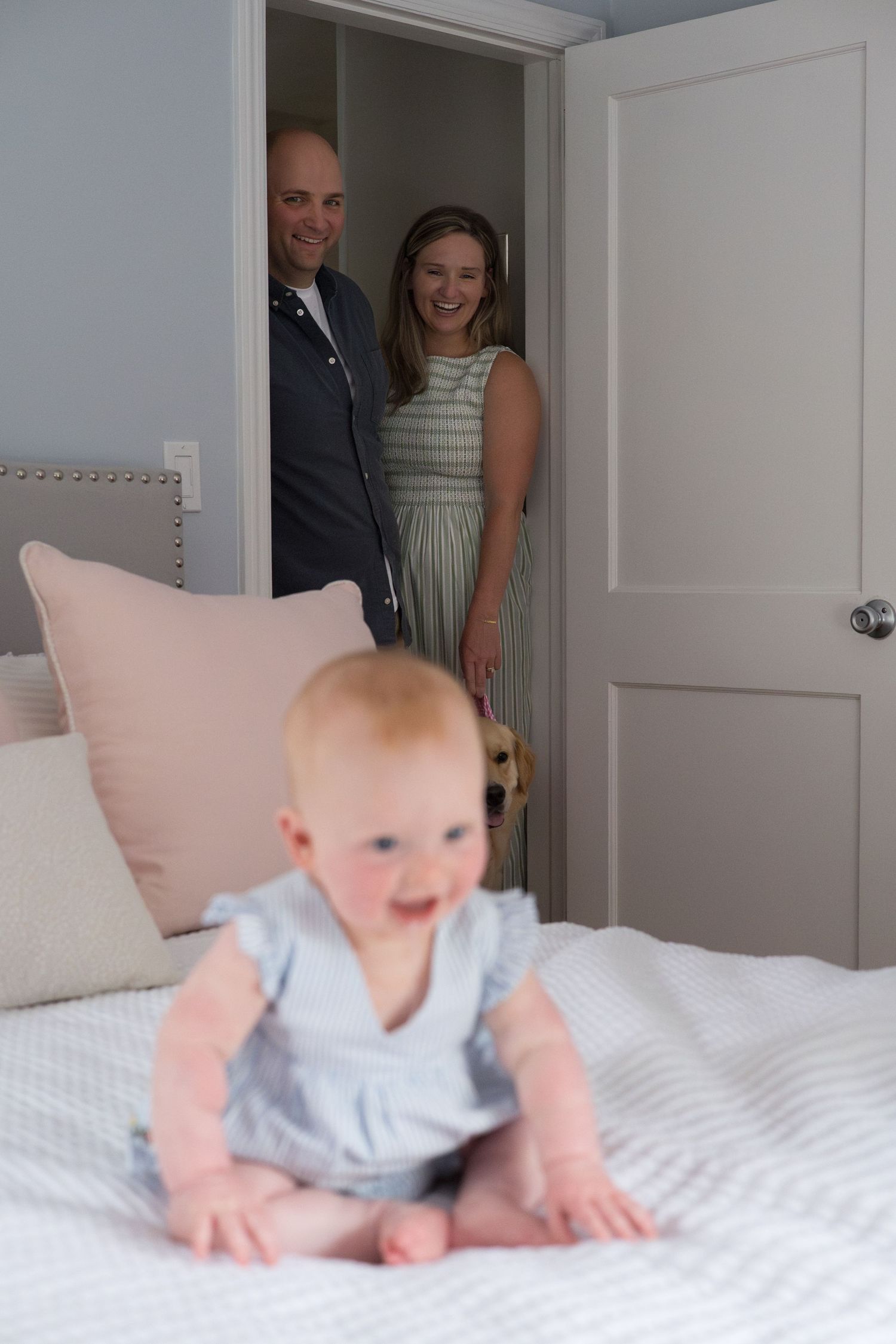 Baby in light blue outfit crawls on white bedding while people watch from doorway in background.