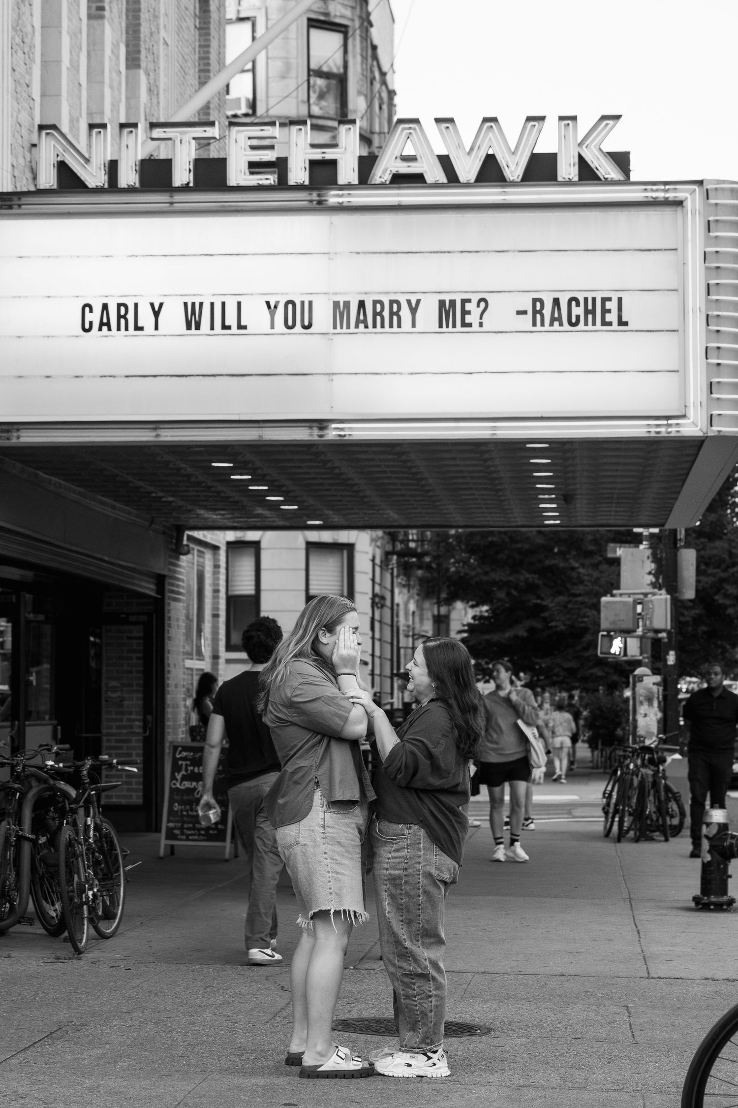 A marriage proposal message is displayed on a theater marquee sign reading 'CARLY WILL YOU MARRY ME? -RACHEL'.