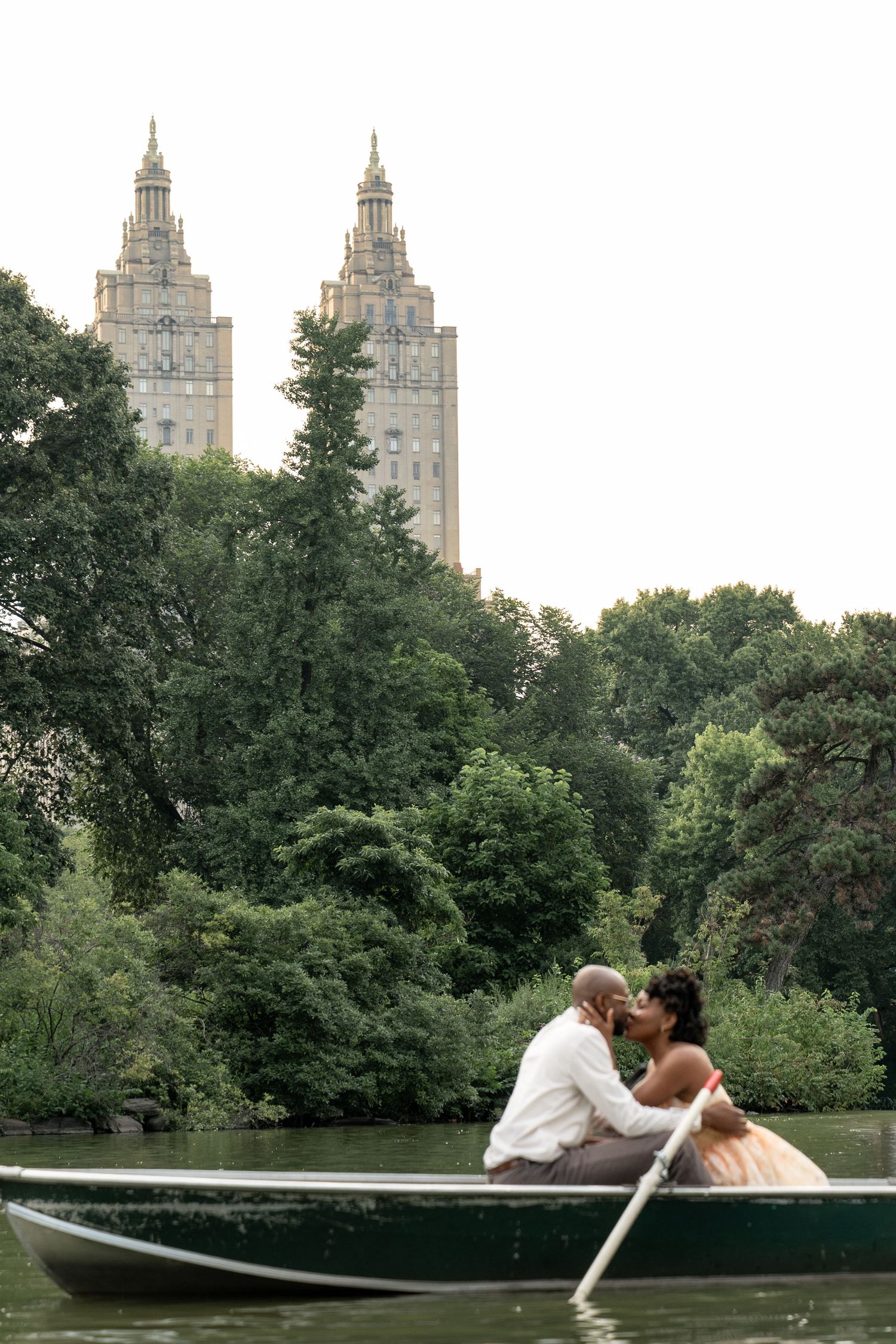 Couple kissing in rowboat on lake with New York City skyline and trees in background.