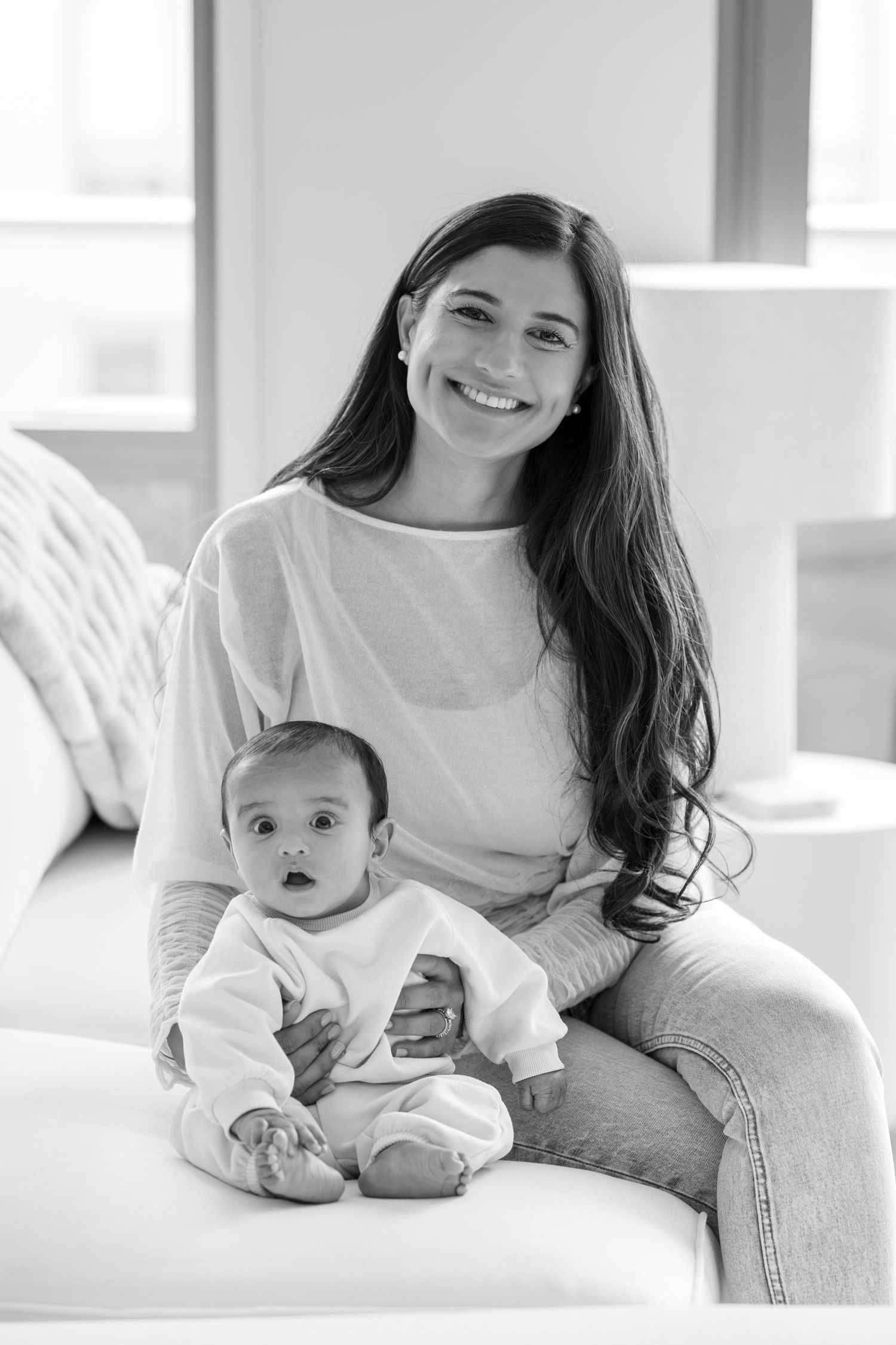 Black and white portrait of smiling brunette mother sitting on sofa with curious baby in white onesie.