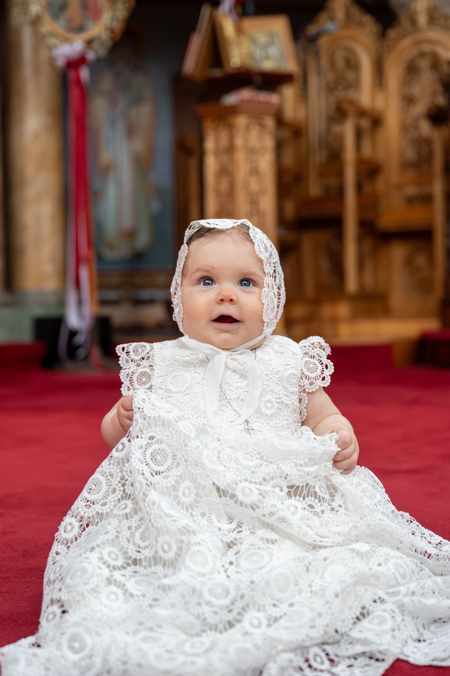 Baby in white lace christening gown and bonnet sitting in a church during baptism ceremony.