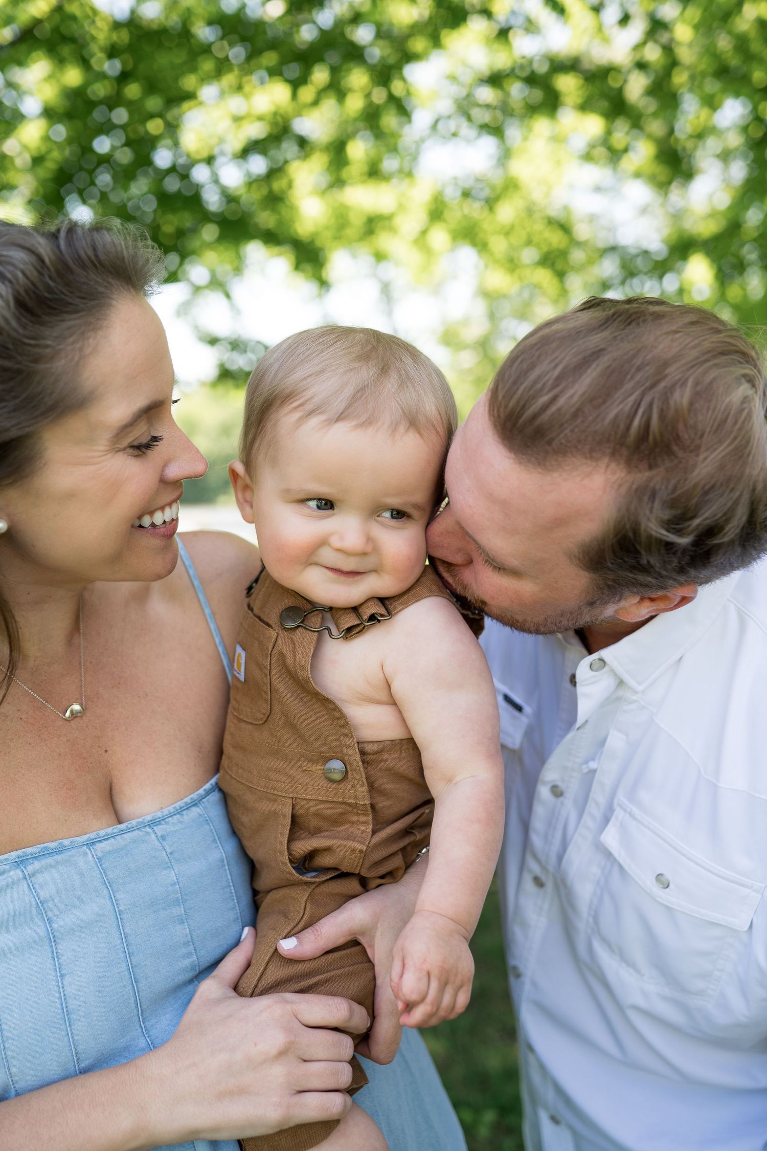 Happy parents kissing and holding their smiling baby outdoors surrounded by lush green trees on a sunny day.