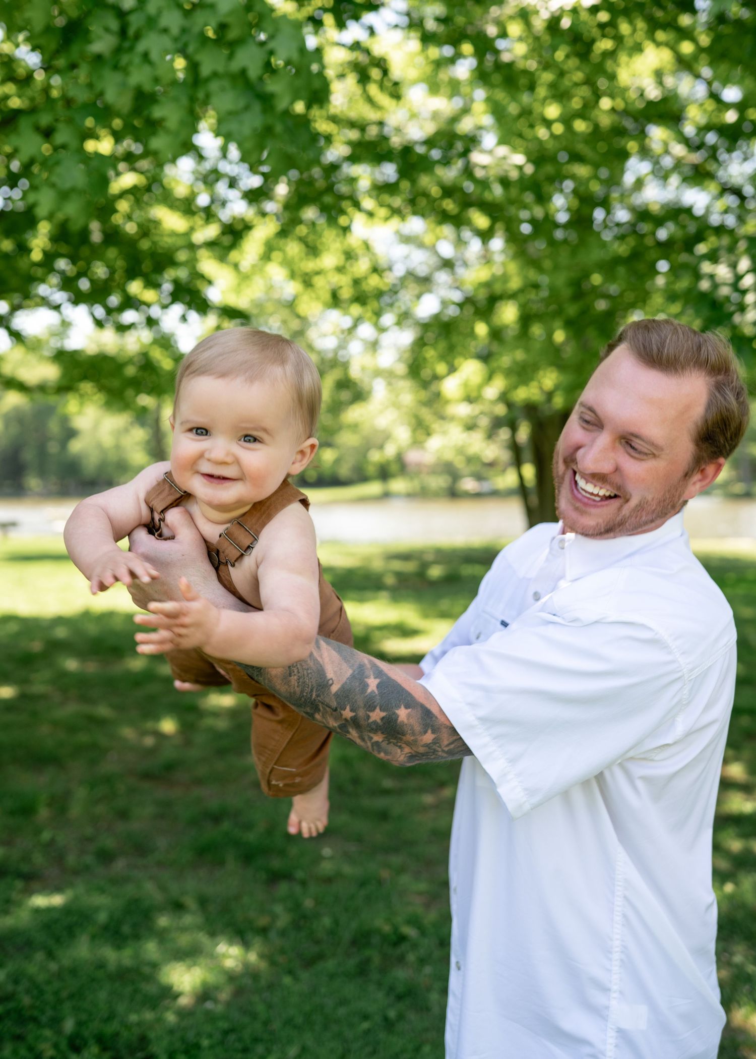 Joyful tattooed father lifting happy baby in brown overalls in lush green park setting.