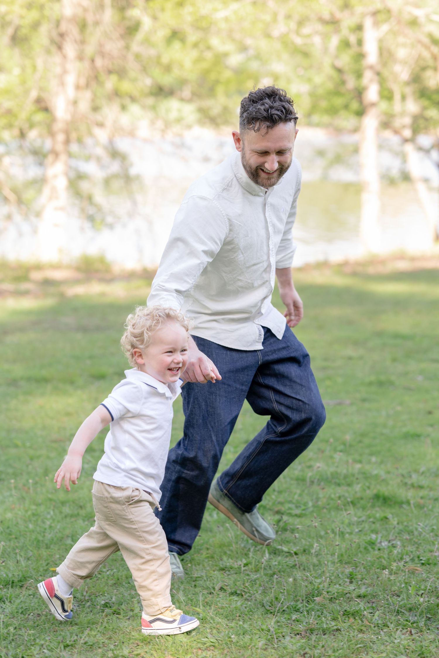 A smiling father chases his laughing toddler son across a green park lawn on a sunny day.