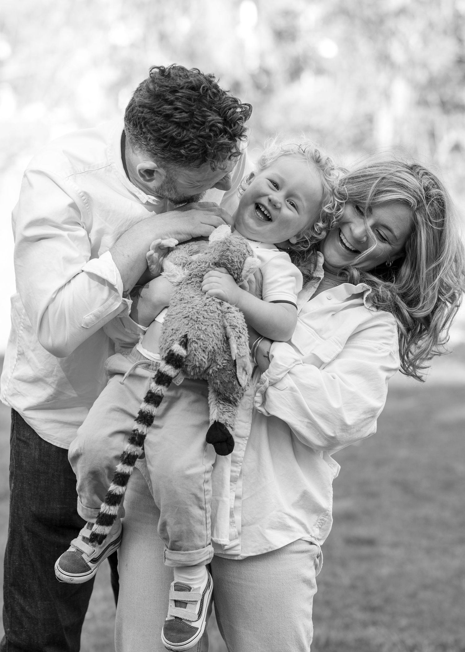 Black and white photo of happy family with parents holding laughing child clutching a ring-tailed lemur outdoors.