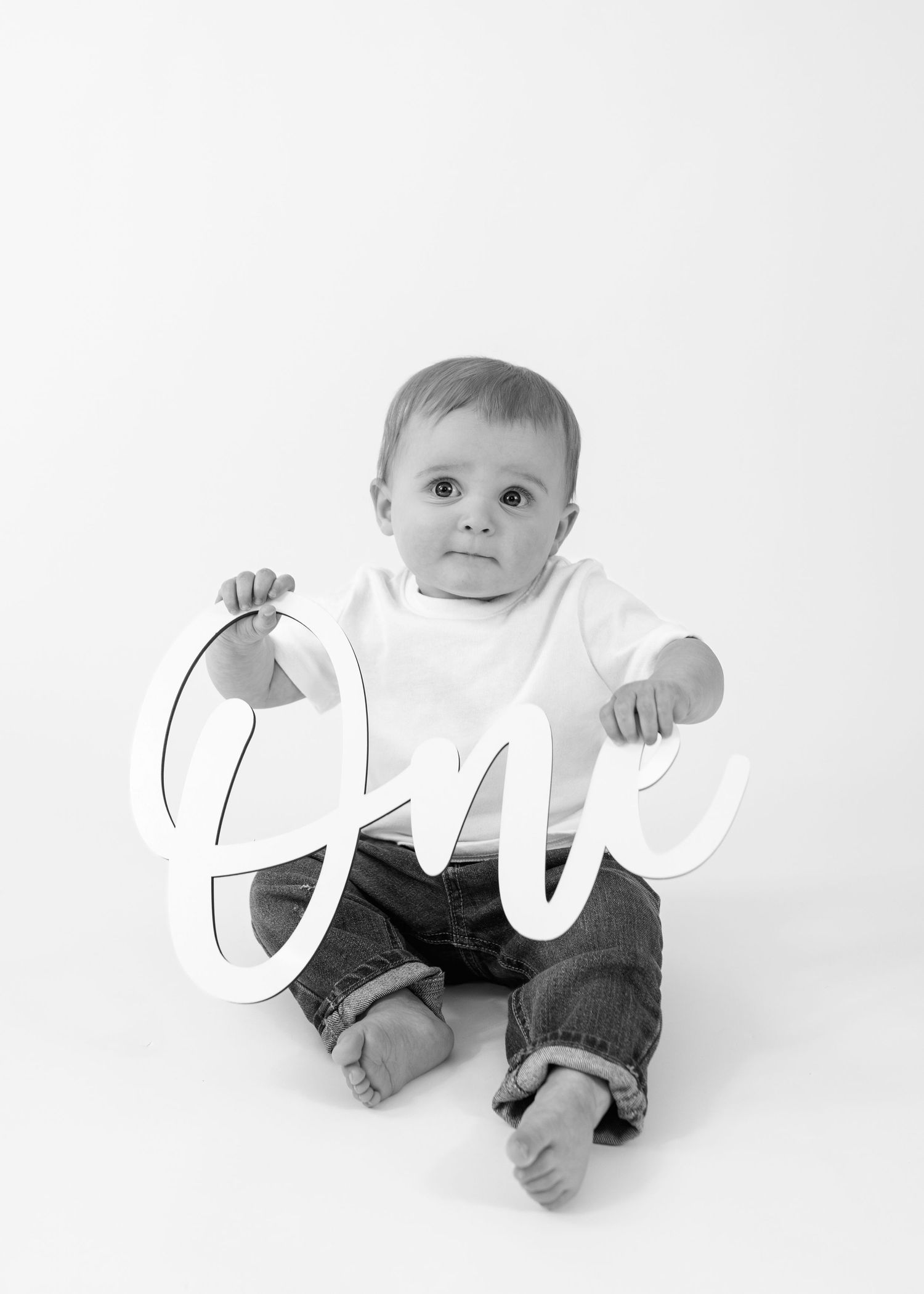 Cute baby boy holding cursive One sign for first birthday photo in black and white.