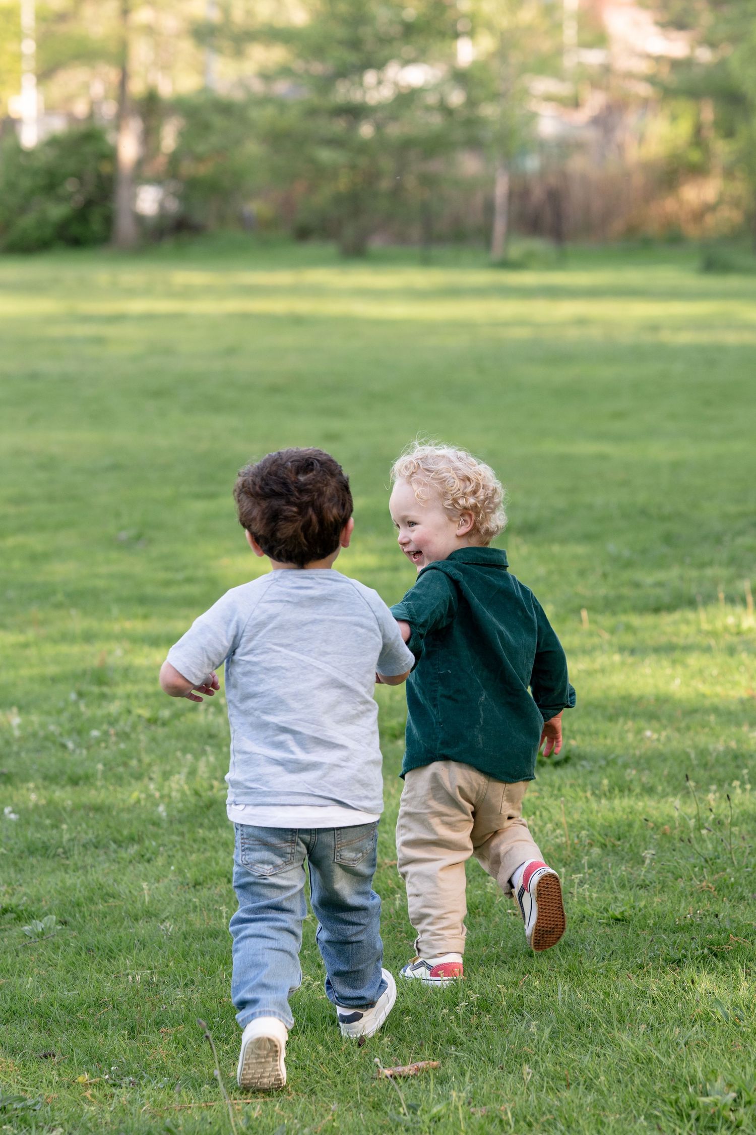 Two toddler boys running together across a lush green park lawn on a sunny day.