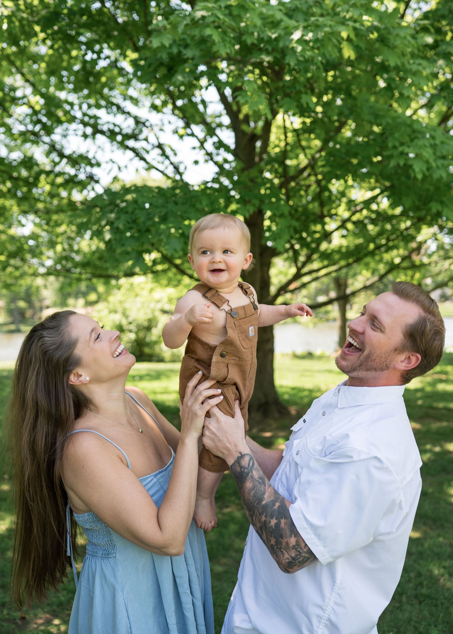 Happy young family laughing outdoors as parents lift smiling baby in brown overalls among lush green trees.